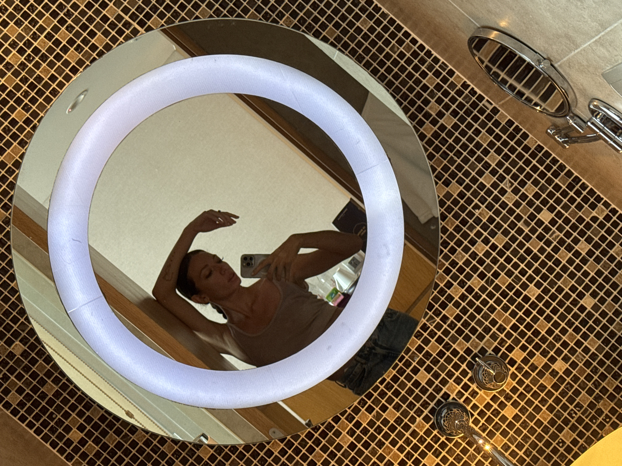 A selfie is taken in a round mirror with a ring light in a bathroom. The mirror reflects a woman with medium-length dark hair and wearing a tank top and blue denim shorts. She is holding a cell phone in her right hand. The bathroom has brown and gold mosaic tile walls and floor. The mirror is affixed to the wall. There is a wall-mounted magnifying mirror with a chrome finish attached to the wall. There is a water faucet. This image shows the bathroom in a hotel in Đà Nẵng, Vietnam.
