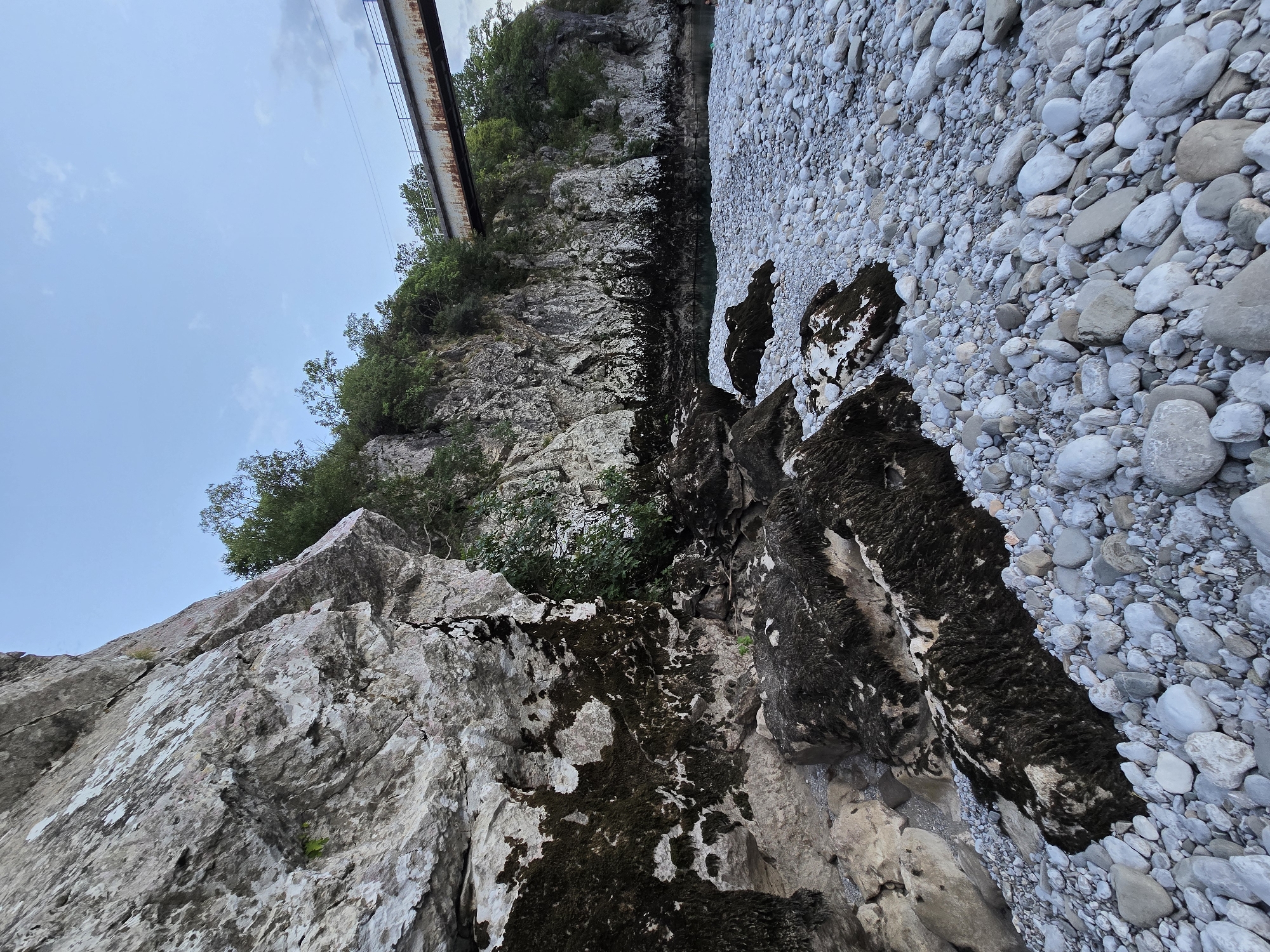 The image depicts a natural landscape scene. In the foreground, a rocky riverbed is covered with numerous smooth, light-colored stones. Some larger rocks, partially covered in dark moss, are scattered throughout the riverbed. To the left, a large, textured rock face rises sharply, with patches of moss and vegetation growing on its surface. Beyond the riverbed, cliffs covered in dense green trees line the bank of a body of water, appearing to be a river. In the background, a long, metallic bridge with railings spans across the top of the scene, crossing over the river. The sky above is a pale blue, with a few wispy clouds. The overall impression is one of a rugged, natural environment under a soft, twilight sky. There are no people visible in the image. This scene is located in Bioče, Montenegro.