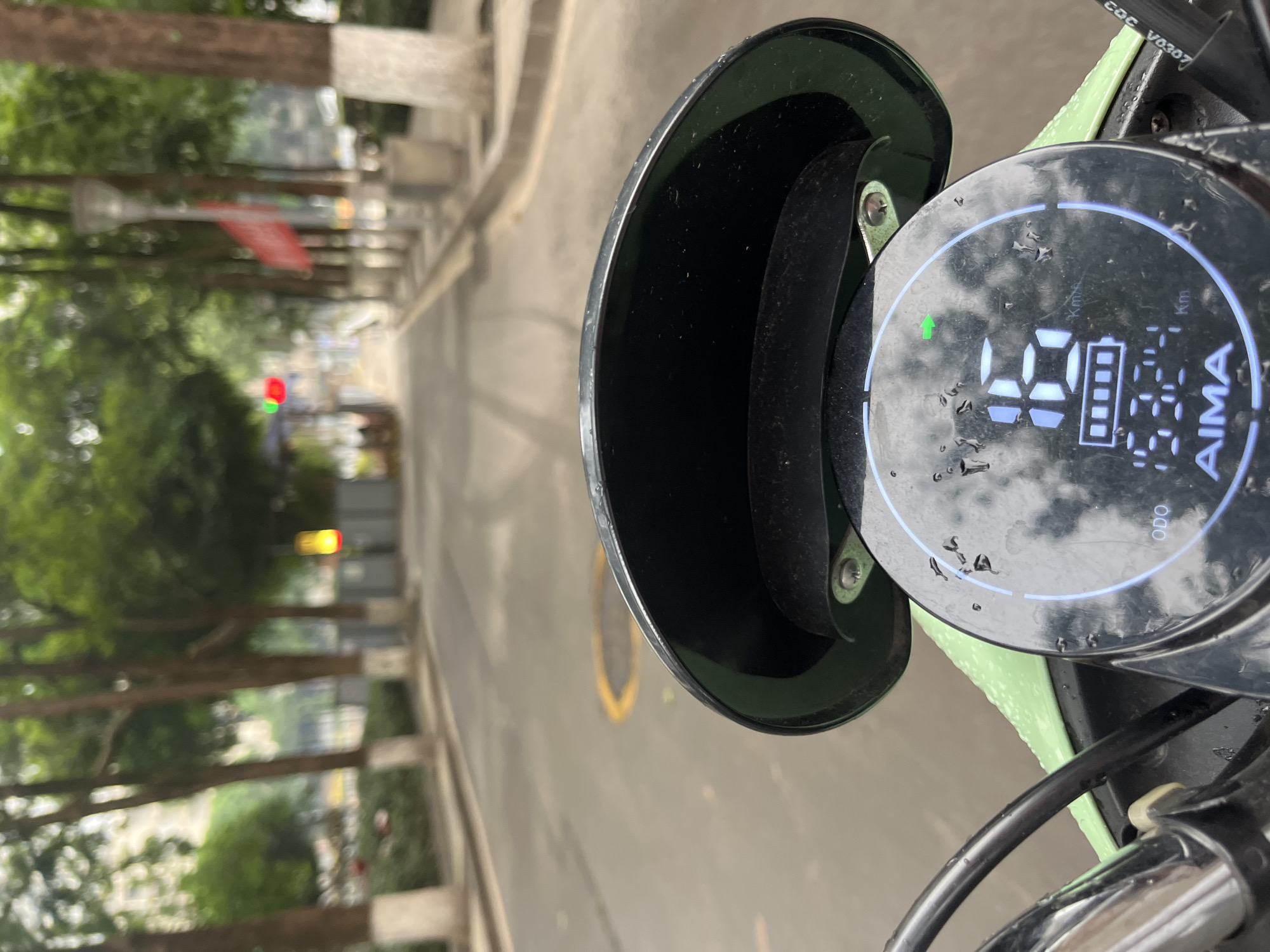 A first-person perspective shot from a motorcycle dashboard, showing a street in Yuzhong District, China. The road is lined with trees and a sidewalk. The dashboard displays a speed of 16 km/h, a full battery, and an odometer reading of 1624 km. The brand name "AIMA" is visible on the dashboard. The scene appears to be during daylight.