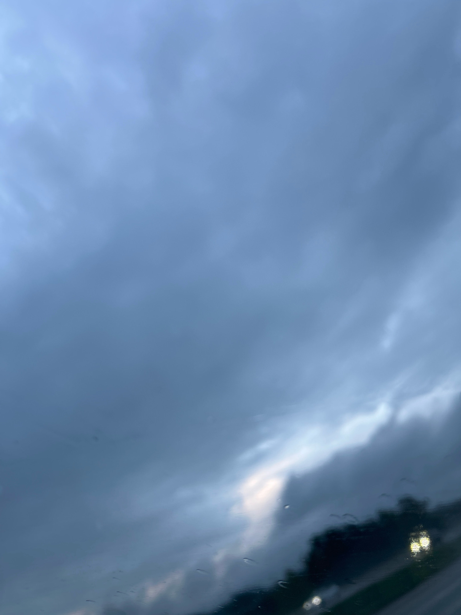 The image depicts a dark, cloudy sky with some lighter patches towards the horizon, suggesting either dawn or dusk, or a break in the clouds. The foreground is out of focus and appears to be a window, possibly of a vehicle, with water droplets visible on the surface, indicating rain. Through the window, the faint silhouettes of trees and streetlights can be seen, indicating an outdoor scene, likely in an urban or suburban area. The overall mood is somber and overcast.