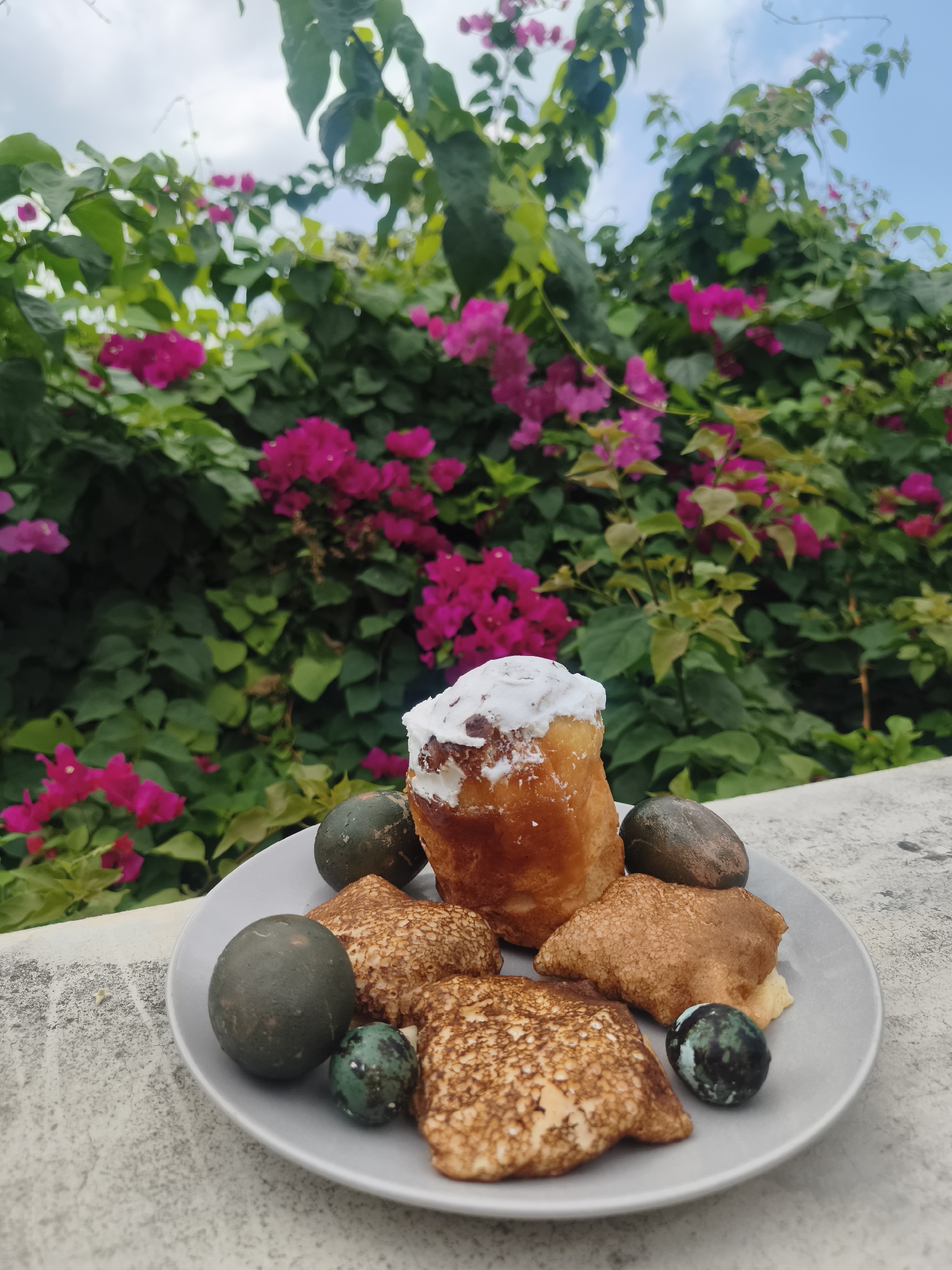 A plate of food sits on a concrete surface, with a backdrop of bougainvillea flowers in full bloom. The food on the plate consists of a tall, cylindrical pastry topped with white frosting, several crepe-like items with a speckled surface, and some dark-colored, round objects that resemble eggs. The location is likely in บ้านบางรักษ์, Thailand, given the likely presence of bougainvillea flowers. No people or interactions are visible in the image.