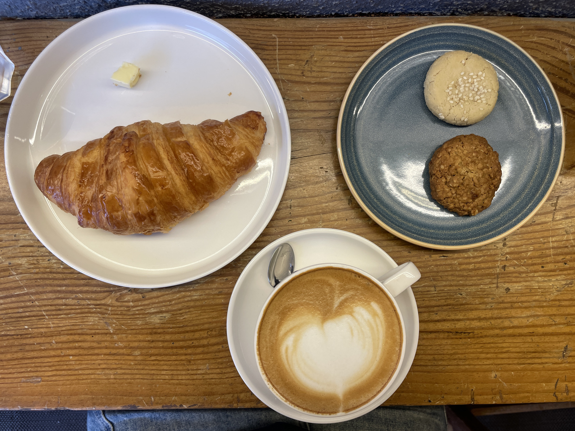 On a wooden table, a breakfast or brunch spread is arranged. A white, round plate holds a croissant and a small pat of butter. Next to it, a blue plate contains two cookies: one light-colored with sesame seeds and one darker, possibly oatmeal cookie. A white cup and saucer with a latte, showing latte art in the shape of a heart, completes the set. The location is Oxel, India, but without further evidence, that information cannot be supported.