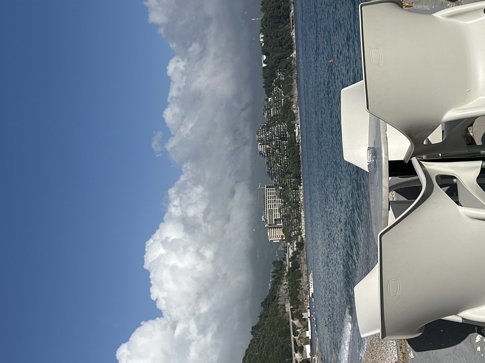 The image shows a coastal view in Budva, Montenegro. The foreground features a stack of white plastic chairs. To the right is the Adriatic Sea, and the coastline is visible, lined with buildings and vegetation. A large, billowing cloud formation occupies the left side of the frame, contrasting with patches of blue sky.