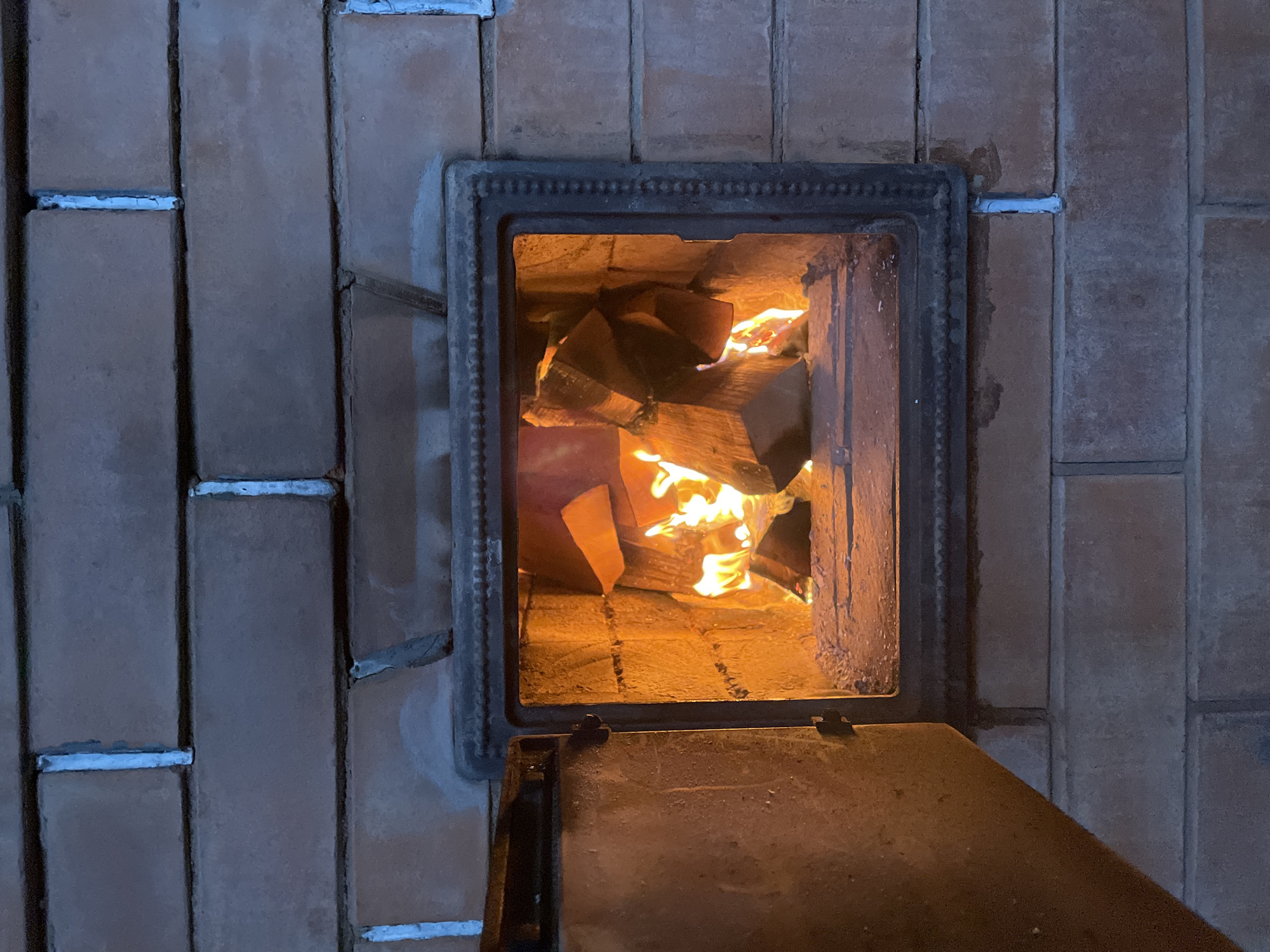 A brick furnace in Sestroretsk, Russia, has a dark metal frame. Inside, burning wood creates a warm glow. The furnace door is partially open, revealing the fire. The bricks surrounding the furnace are laid vertically, with white mortar visible between them.