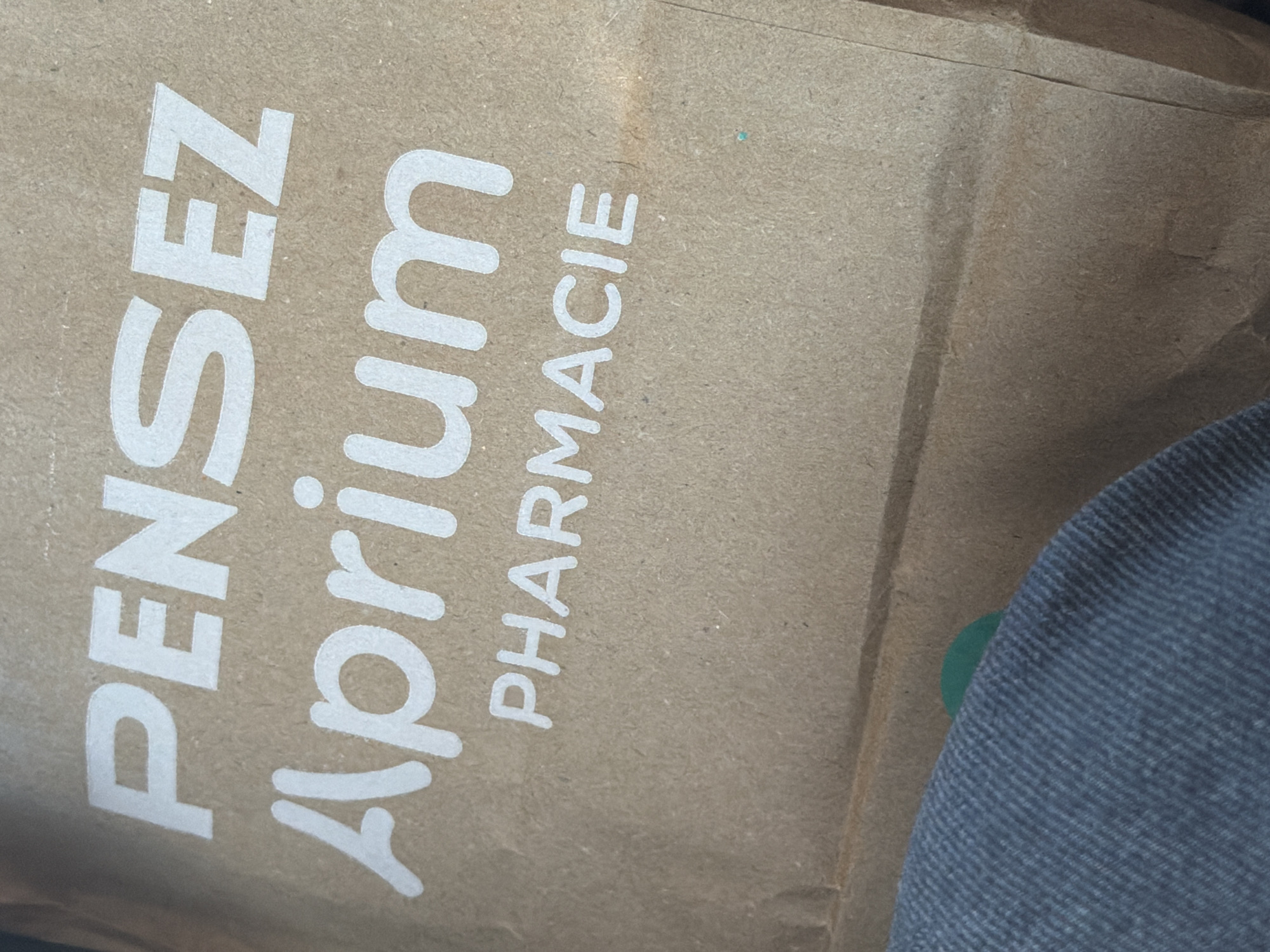 The image shows a close-up of a brown paper shopping bag. White text on the bag reads "PENSEZ Aprium PHARMACIE". The bag is made of recycled material and has a slightly textured surface. The bottom left of the image shows a portion of dark blue denim fabric, possibly clothing. A small, out-of-focus green circle is visible on the bag, near the denim. The lighting suggests an indoor environment, possibly a store or a home. The location is not visible or inferable from the image.