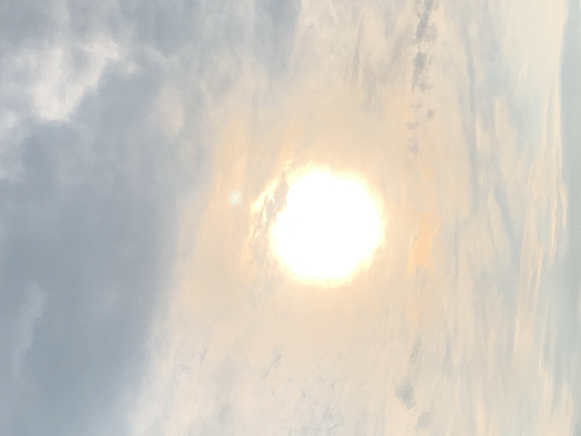 The image depicts a bright sun, slightly obscured by clouds, in a sky that transitions from a pale orange near the sun to a soft blue and grey further away. Thin, wispy clouds are scattered throughout the sky, with some denser, darker clouds present in the upper portion. There are no people, subjects, objects, or actions visible in the image. The scene appears to be an outdoor environment, likely during the daytime. The provided information regarding the city of Kolomyia, Ukraine, is not visually discernible from the image itself.