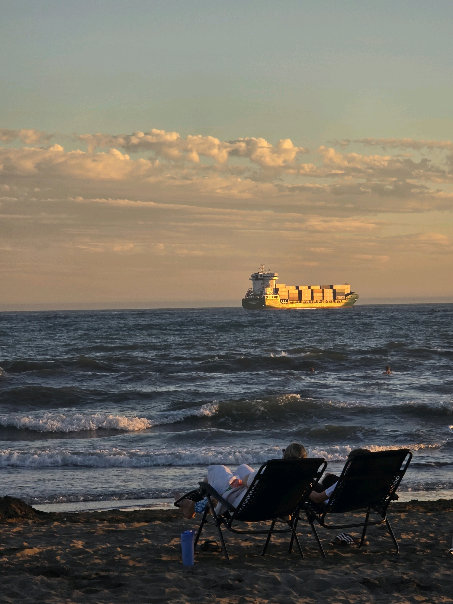 The image depicts a tranquil beach scene at sunset, overlooking the Mediterranean Sea. In the foreground, two figures recline in lounge chairs on the sand, facing the water. The sun, low on the horizon, casts a warm, golden light across the scene, illuminating the clouds in soft hues of orange and pink. The sea is moderately choppy, with waves breaking gently on the shore. Further out, a cargo ship is visible, its deck filled with stacked containers, suggesting a busy shipping lane. The overall atmosphere is one of peaceful relaxation and natural beauty. The location is identified as Málaga, Spain, a coastal city known for its picturesque beaches and vibrant maritime activity.