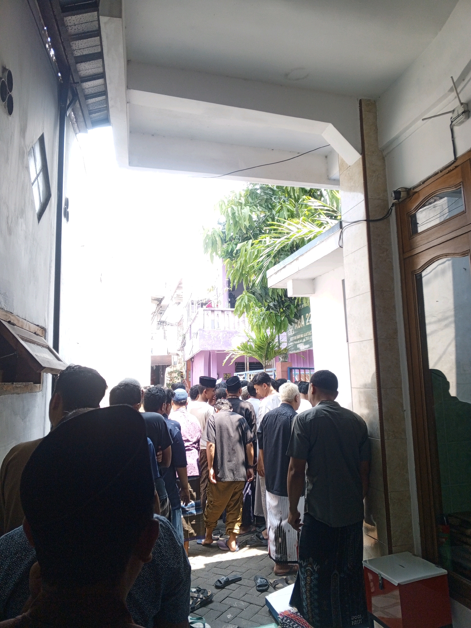 A group of men, dressed in traditional Islamic attire including peci hats and sarongs, are gathered outside a building, possibly for a prayer or ceremony. The scene appears to be in a narrow alleyway, with one side of the alley being a grey concrete wall and the other side a building with a wooden door and glass. Sunlight is bright, casting strong shadows, and suggesting an outdoor, daytime setting. There's a red cooler box on the ground near the wooden door. A sign with Indonesian text is visible on the building in the background. The overall impression is one of a community gathering.