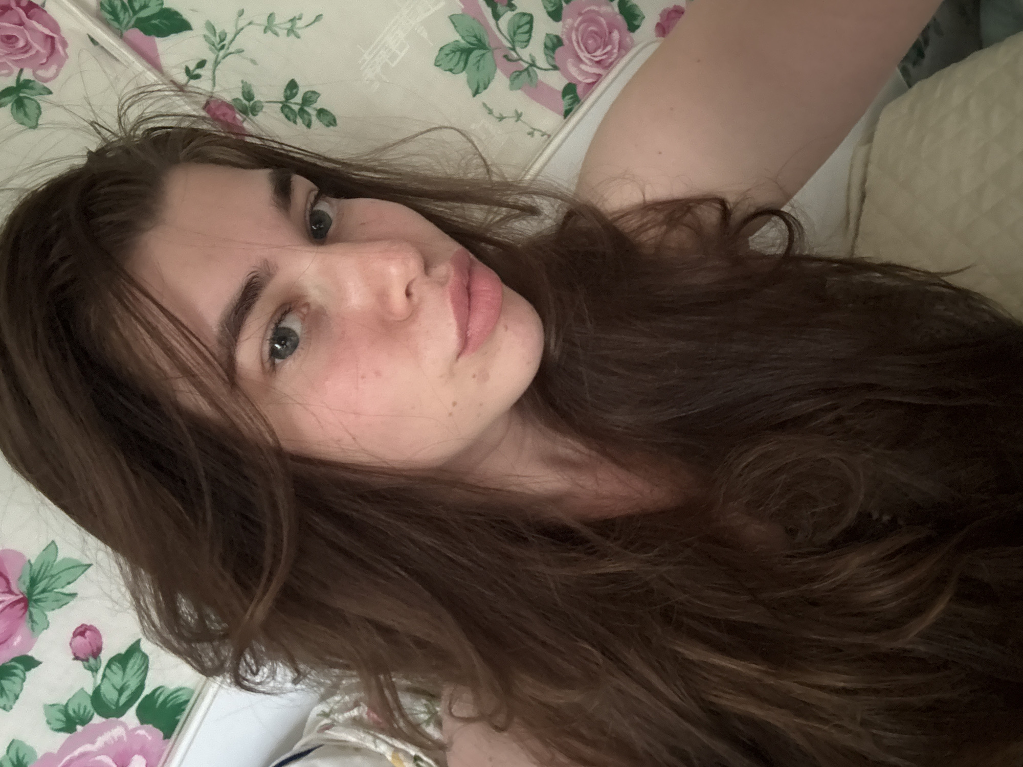 A young woman with long, wavy brown hair is captured in a close-up selfie. She is lying down against a floral-patterned background, possibly bedding or wallpaper, featuring pink roses and green leaves. Her skin is fair with a slight flush on her cheeks and a smattering of freckles across her nose and cheeks. Her eyes are a striking blue-green, and her lips are full and painted a soft pink. The lighting suggests a natural, indoor setting. The scene does not contain any information that could indicate a specific location within the city of Мещёры, Russia.