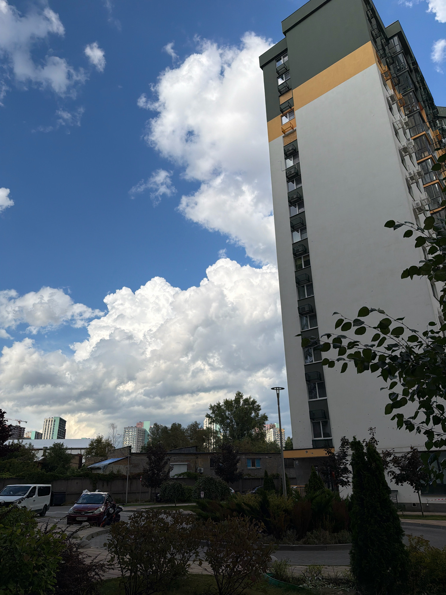 The image captures a street-level view of a modern, multi-story residential building under a partly cloudy sky in Kyiv, Ukraine. The building, predominantly white with a dark green top section and a horizontal ochre band, dominates the right side of the frame. It features numerous windows and what appear to be external air conditioning units or decorative elements on balconies. In the foreground and mid-ground, there are various trees and bushes, some appearing well-maintained. A paved road and sidewalk are visible, with a white van and a red sedan parked on the side of the road. In the distance, other residential buildings and construction cranes are visible, suggesting an ongoing urban development. The sky is a vibrant blue with large, fluffy white clouds. The overall impression is of a contemporary urban environment on a clear day.