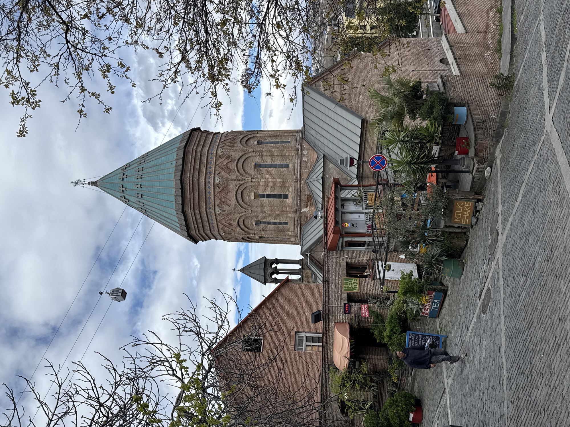 The image shows the Narikala Fortress in Tbilisi, Georgia, viewed from below. The tall brick tower with a conical turquoise roof dominates the frame, reaching into a cloudy sky. Bare tree branches with emerging leaves frame the top and left side of the image. A smaller tower with a dark conical roof stands beside the main tower.
Buildings constructed from brick and other materials, adorned with signs such as "RED WINE," "MILK SHAKE," and "HERBAL TEA," suggest the presence of shops or restaurants. A "No Stopping" sign is also visible. A person stands on a cobblestone street, likely a pedestrian observing the scene. A cable car car is seen hanging from a cable overhead.