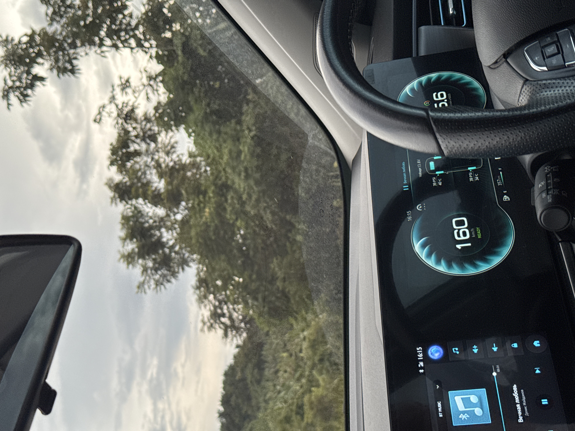 A car's dashboard display shows the car is traveling at 160 km/h. The time is 16:15. The car's voltage is 13.8V. Tire pressure is 39 PSI for the front tires and 34 PSI for the rear tires. The outside temperature is 40°C for the front tires and 35°C for the rear tires. The car has traveled 357 km. The car stereo is playing a song titled "Вечная любовь" by Денис Майданов. The view through the windshield shows trees and a cloudy sky, suggesting a rural or natural setting. The location is Plai Phraya, Thailand.