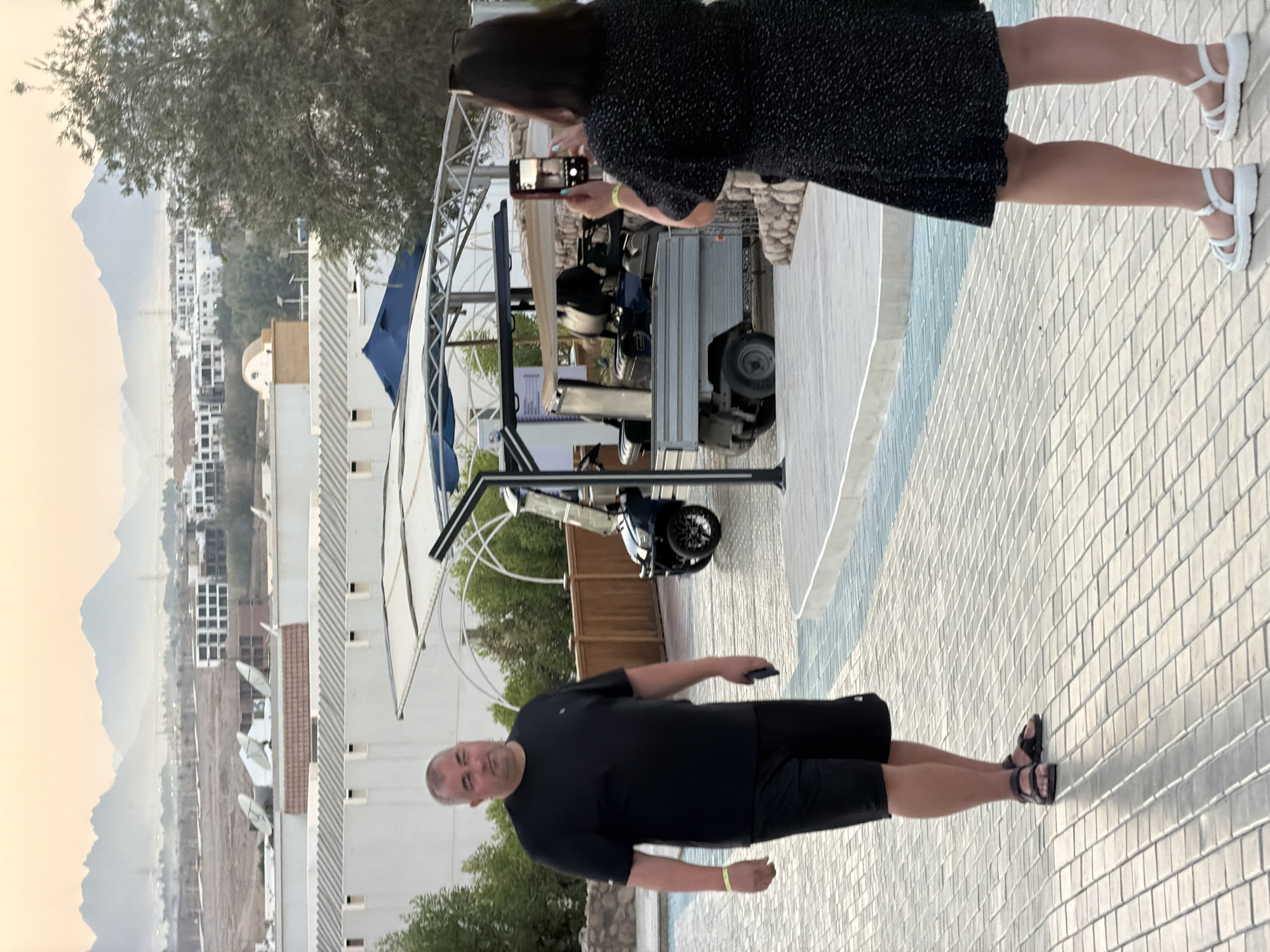 In Sharm El Sheikh, Egypt, a man in a black t-shirt and shorts stands on a tiled pathway, looking to his left. He is holding a dark mobile phone in his right hand and wears dark sandals. A woman in a black polka-dotted dress is taking a photo of him with her mobile phone, which she holds with both hands. She is wearing white sandals. In the background, a golf cart is parked under a covered structure. The environment appears to be an outdoor resort or public area with buildings and vegetation visible, and mountains in the distance under a hazy sky.