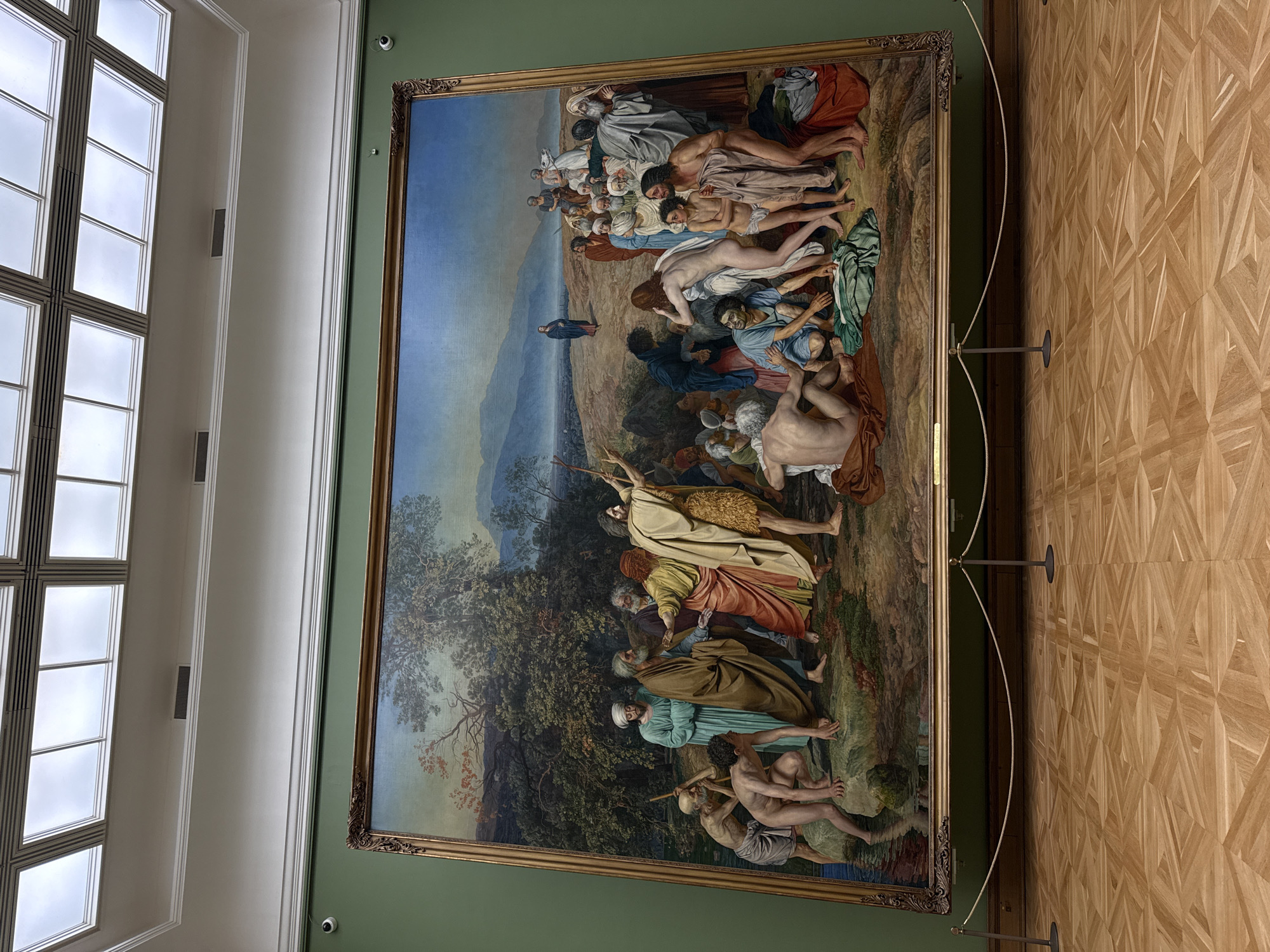 The image shows a large painting in a museum setting. The painting is a historical scene, depicting a large group of people in various states of distress and action. The upper portion of the painting features figures in a mountainous landscape, with a central figure seemingly isolated in the midground. The lower portion depicts a gathering of individuals in what appears to be a wooded area.
The painting is framed in ornate gold and is displayed against a green wall. The museum setting is indicated by the parquet floor and a support cable for the painting. Based on the art style and common displays, this could possibly be located in the Pushkin State Museum of Fine Arts in Moscow, Russia. The lighting in the room is from natural light coming through a large, multi-paned window that can be seen on the left. A security camera is visible on the wall near the painting.