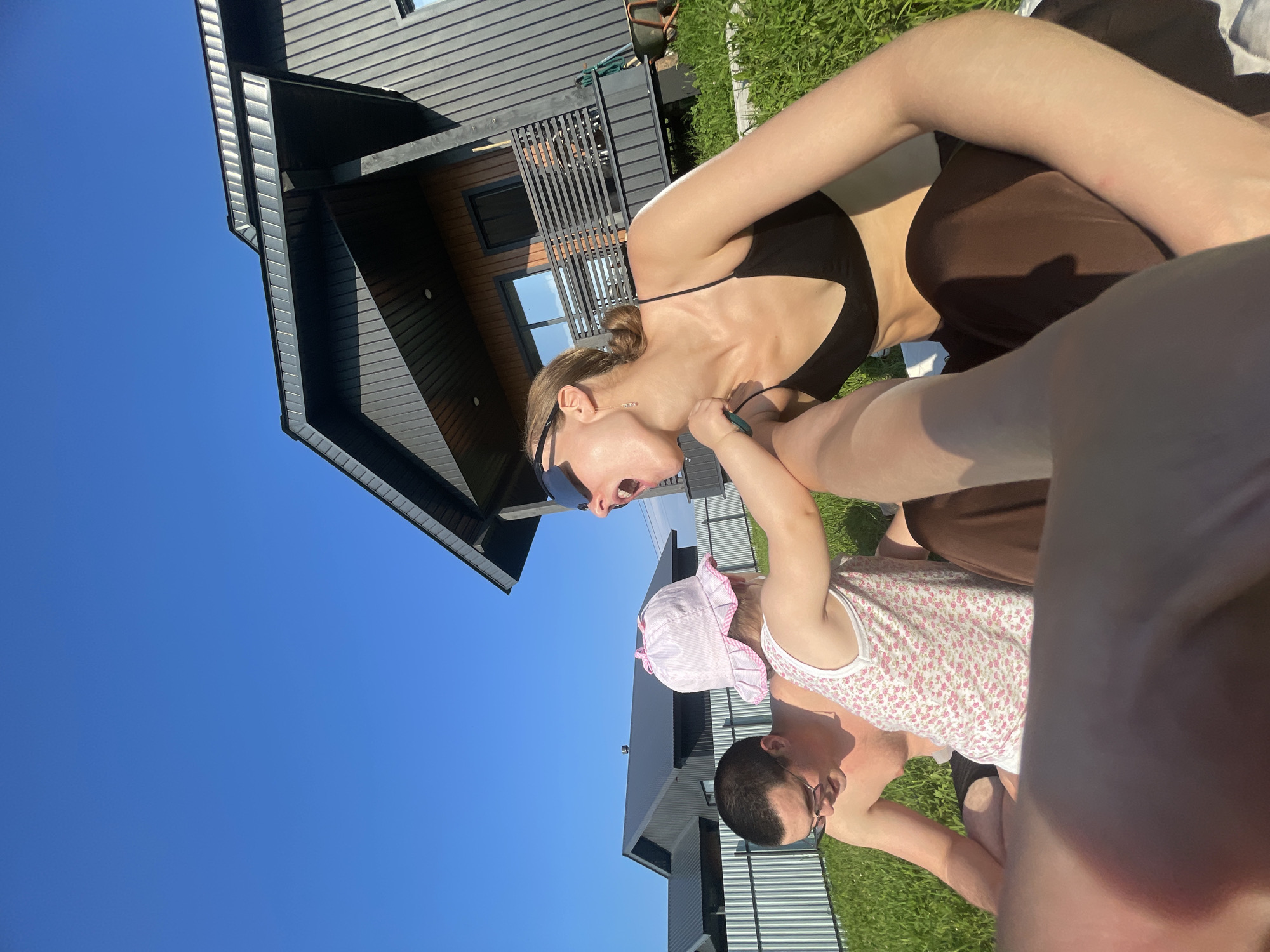 In this overhead, low-angle shot, a family enjoys a sunny day in a grassy yard near a modern house. The sky is a vibrant, cloudless blue, indicating clear weather. The house features dark siding and a contemporary design with large windows.
The main subjects are two adults and a baby. A woman in a black bikini top and brown bottoms is leaning forward, reaching towards the baby. She is wearing sunglasses. The baby, dressed in a pink bonnet and a floral tank top, is being held by another adult, a man wearing glasses. The man is also leaning forward, looking at the baby. The interaction suggests a playful moment between the family members.
The sun is high in the sky, casting distinct shadows, suggesting it is midday. The lush green grass indicates a warm season. The overall mood appears relaxed and joyful, with the family enjoying outdoor time together. No visible text is present in the image. The setting is a residential area, likely a backyard in Подшивалово, Russia.
