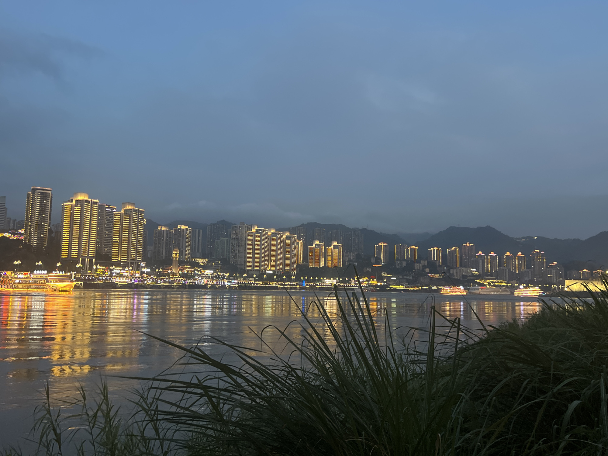 The image depicts a nighttime cityscape of Jiangbei Cheng Street, China, viewed across a body of water. The skyline is dominated by numerous illuminated high-rise buildings, their warm yellow lights reflecting on the water's surface. Several boats are visible on the river, also lit up, adding to the vibrant nocturnal scene. In the foreground, lush green reeds or tall grass create a natural border, partially obscuring the view of the water and cityscape. The sky is a deep, dusky blue, with some faint cloud cover, suggesting it is either dusk or pre-dawn. The overall atmosphere is serene and picturesque, highlighting the urban development and the natural elements of the location.