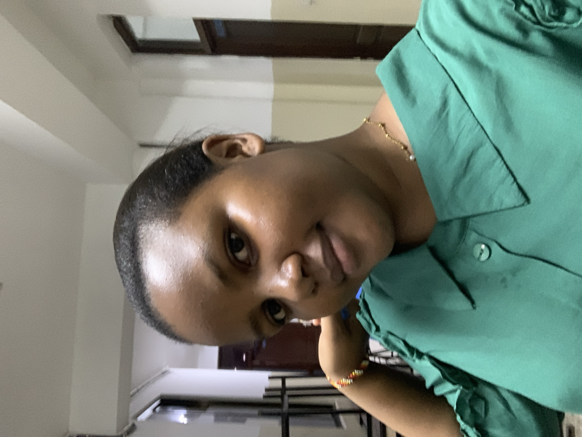 A person with dark hair is wearing a green shirt and a gold necklace. They are wearing a colorful beaded bracelet on their left wrist. They appear to be inside a building with white walls and a dark wood door. There are metal bars in the background. The location is not possible to determine based on the image provided.