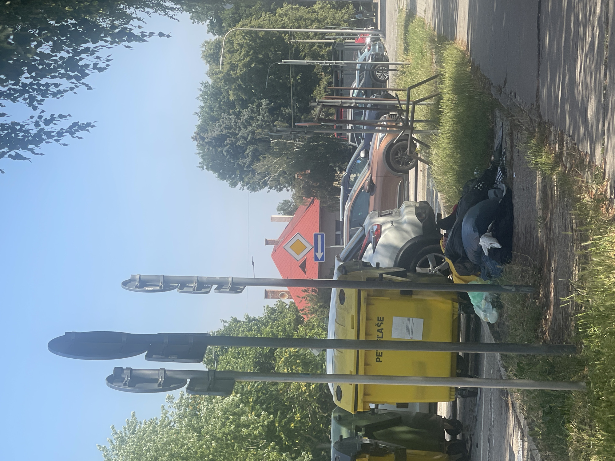 In an outdoor urban setting, a yellow recycling bin labeled "PET | PLASE" is positioned next to other bins, including a green one. Piled on the ground next to the yellow bin are several trash bags and bundled items, including a white glove. Several cars are parked in the background, including a brown SUV and a silver car. Trees and foliage line the street, and a street light is visible. A red-roofed building with a diamond-shaped yellow sign and a blue arrow pointing left is in the distance. The scene appears to be a street or parking area under a clear blue sky.