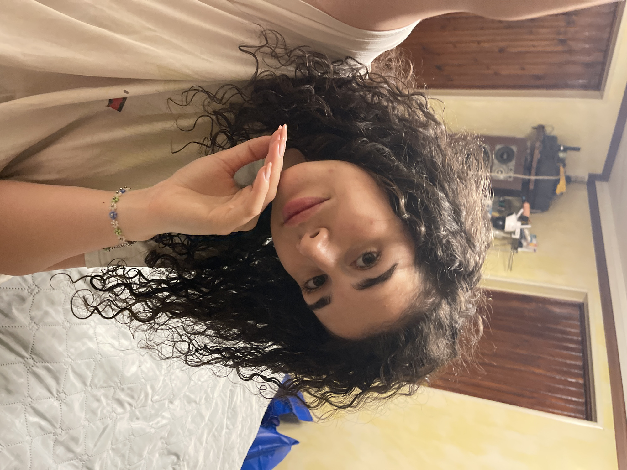 A young woman with curly dark hair, wearing a white t-shirt and a bracelet with small blue and green beads, is posing for a selfie. She is leaning her head on her hand, her arm resting on her chest. The background shows a bedroom with wooden doors, yellow walls, and a white quilted bedspread. The image is taken indoors, possibly in an apartment in Sofia, Bulgaria.