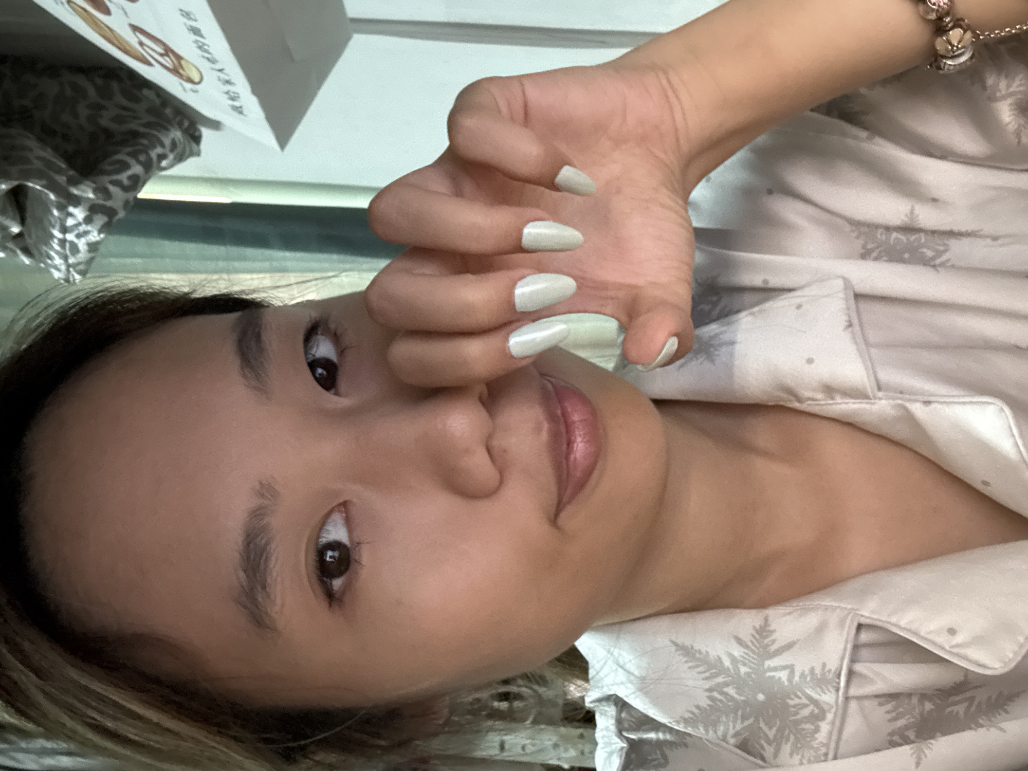 A woman with light brown hair and skin is facing the camera. She has brown eyes and is wearing a light-colored pajama top with a snowflake pattern. Her hand is raised in front of her face, showing light green, almond-shaped nails. A charm bracelet is visible on her wrist. A shopping bag and part of a patterned pillow are in the background. The location is likely inside a residence in Nan'an District, China, as inferred from the context.