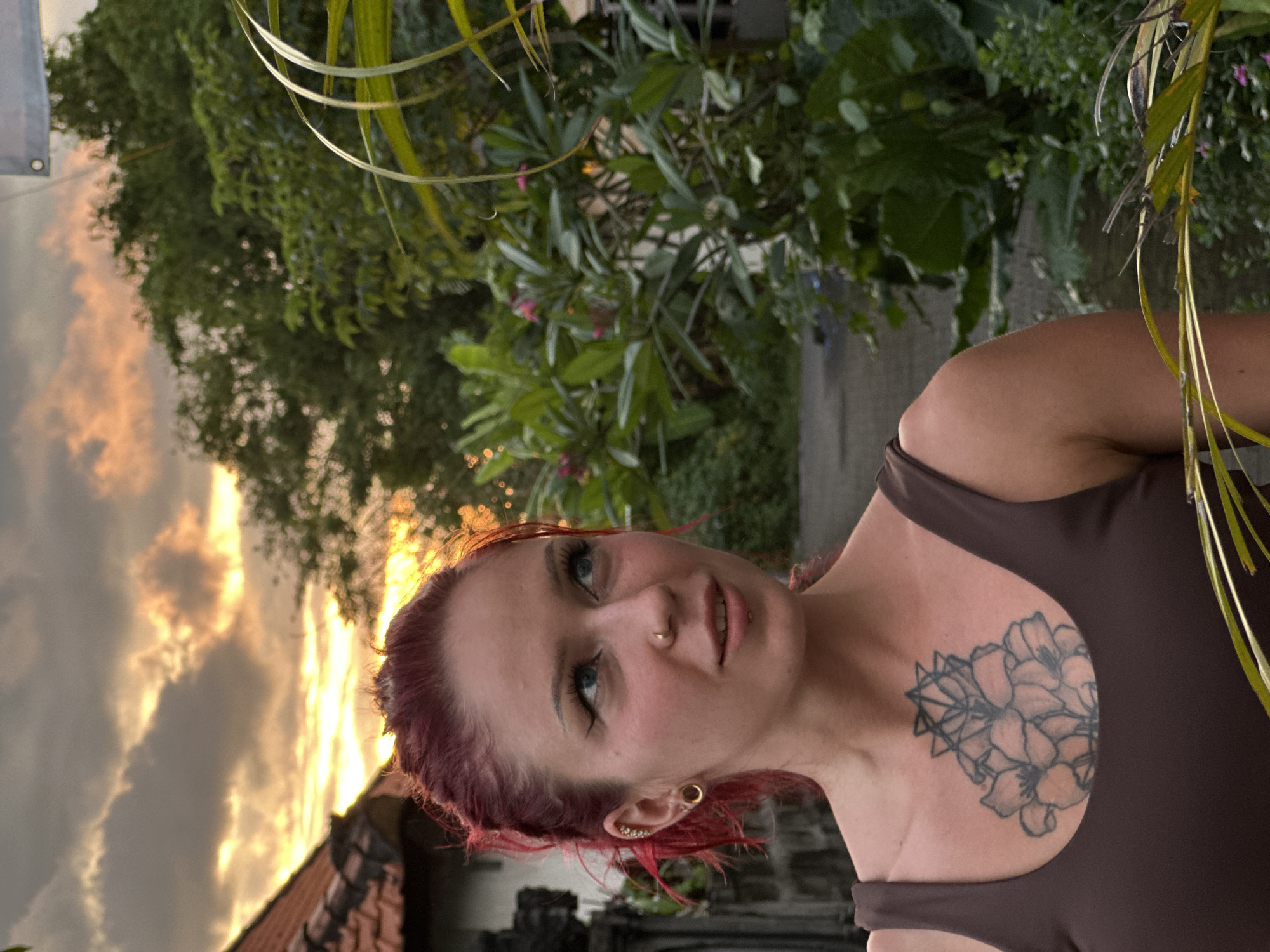 A woman with reddish hair and blue eyes is pictured in a partially obscured outdoor setting, possibly in Dalung, Indonesia. She is wearing a brown sleeveless top, and a floral tattoo is visible on her upper chest. The background shows foliage and a partly cloudy sky with what appears to be the sun setting, casting a warm light. The overall scene suggests a natural, potentially tropical environment. A building is visible on the left side of the image.