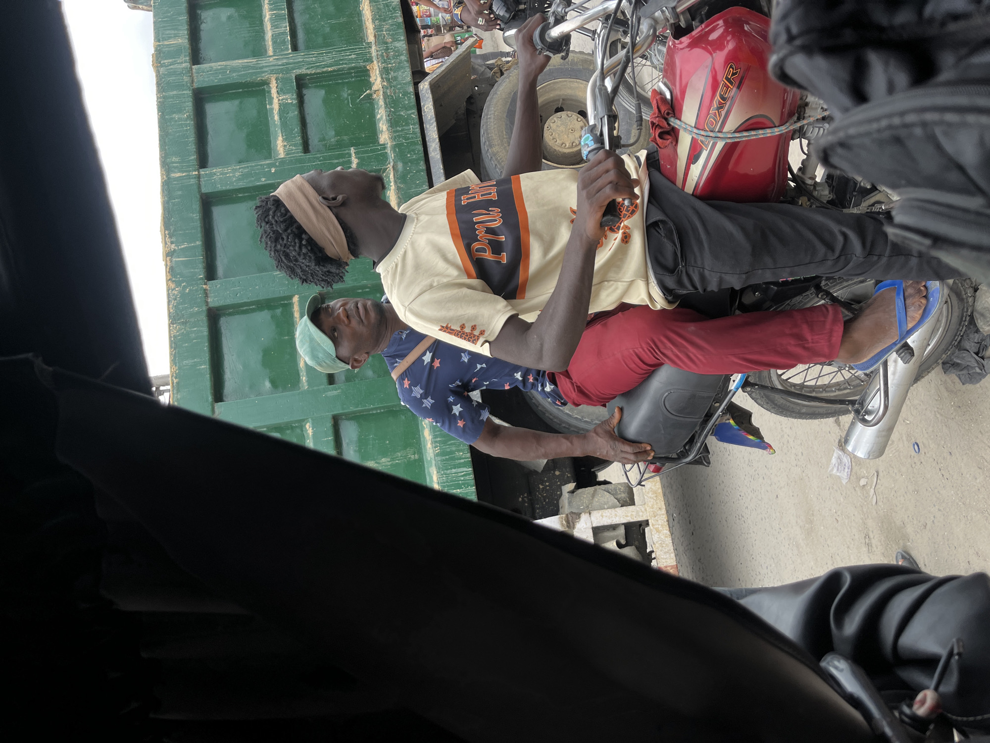 The image shows two men on a motorcycle stopped next to a green truck. The man in front wears a tan shirt with a black stripe and "Prv Ht" on it, black pants, a tan headband, and blue flip-flops. He holds the motorcycle handlebars. The man behind him wears a blue shirt with white stars, red pants, and a green cap. He is holding onto the motorcycle seat. The motorcycle has a red gas tank labeled "BOXER." The scene appears to be on a paved road next to a large green truck. Based on common features in the city, it is likely this scene occurred in Lekki, Nigeria.