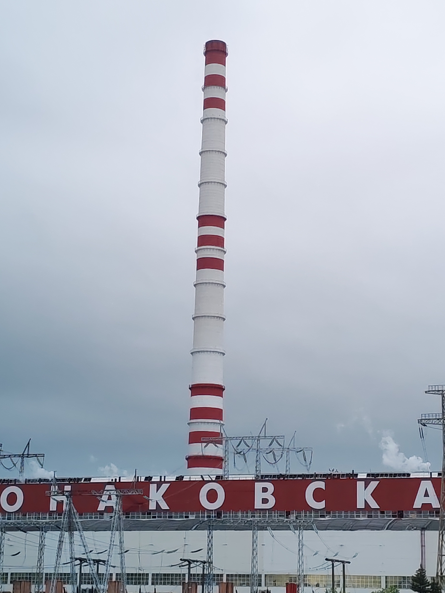 A tall, red-and-white striped smokestack dominates the center of the image. It is part of the Konakovska Thermal Power Plant, with the name "KOHAKOBCKA" visible on the building below. The power plant appears to be a large, industrial structure with numerous windows and overhead electrical lines. The sky is overcast and gray. The location is Konakovo, Russia.