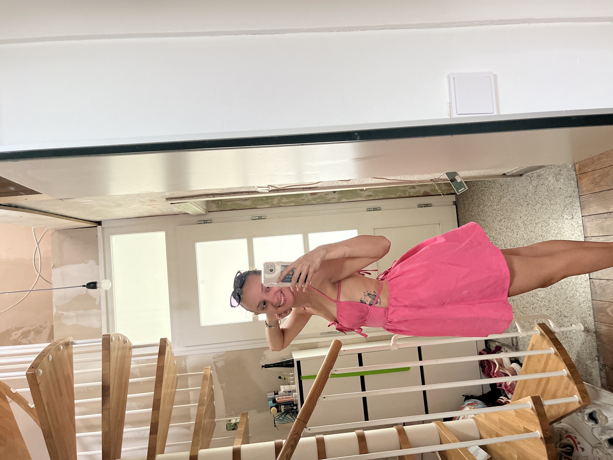 A young woman with blonde hair is taking a selfie in a mirror. She is wearing a bright pink dress with spaghetti straps and a cutout on her torso, revealing a butterfly tattoo on her chest. She is smiling at the camera and wearing sunglasses on her head. The background appears to be a room under renovation, with exposed walls and scaffolding. To the left, a wooden staircase with white railings is visible. To the right, there is a white cabinet with shelves holding various items, including bottles and shoes. A closed white door with glass panes is also in the background. The lighting suggests it is daytime, possibly indoors with artificial light. The overall mood is casual and cheerful. The image is a vertical orientation.