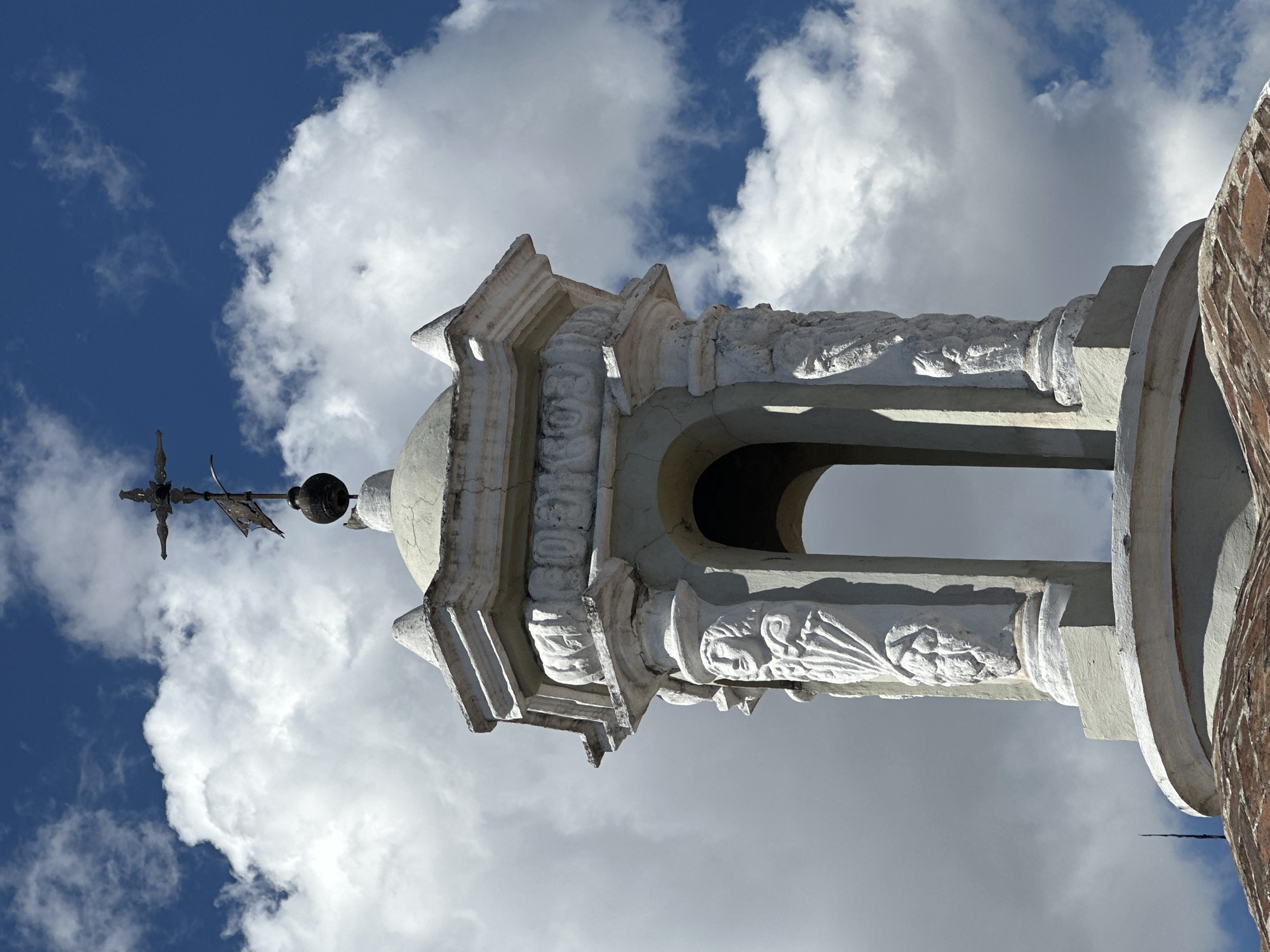 The image shows the upper portion of a bell tower or belfry, likely part of a church or cathedral in Puebla, Mexico. The structure is made of white stone or plaster, with ornate carvings on its columns and decorative elements. Each of the visible columns features a relief of a face, possibly a saint or an allegorical figure, with detailed drapery. Above these columns, there is an octagonal structure with decorative moldings and a small dome. Crowning the structure is a weather vane topped with a cross, suggesting it's a religious building. The sky in the background is a vibrant blue with fluffy white clouds, indicating a bright, clear day. The lower edge of the image reveals a part of a reddish-tiled roof, likely belonging to the same building. The architectural style suggests a historical building, possibly colonial or baroque, common in Puebla.