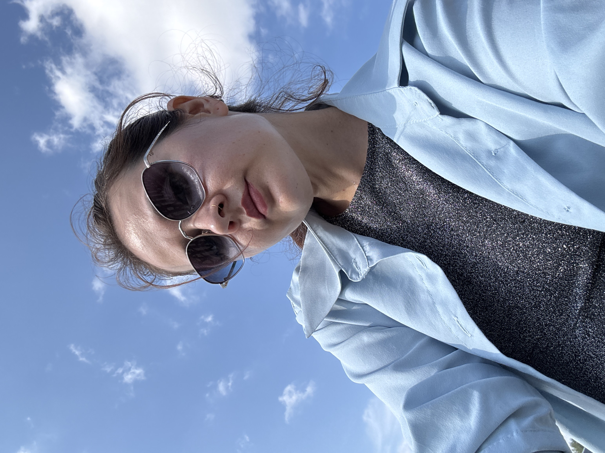 An upward-angle, medium shot captures a young woman looking slightly down, her face framed by the bright blue sky with scattered white clouds. She wears oversized, silver-rimmed sunglasses with dark lenses and a light blue open trench coat over a dark, shimmering top. Her dark hair is pulled back, with wisps escaping in the wind. A small nose ring is visible. The image appears to be taken outdoors on a sunny day. There is no specific indication of the city of Naberezhnye Chelny, Russia.