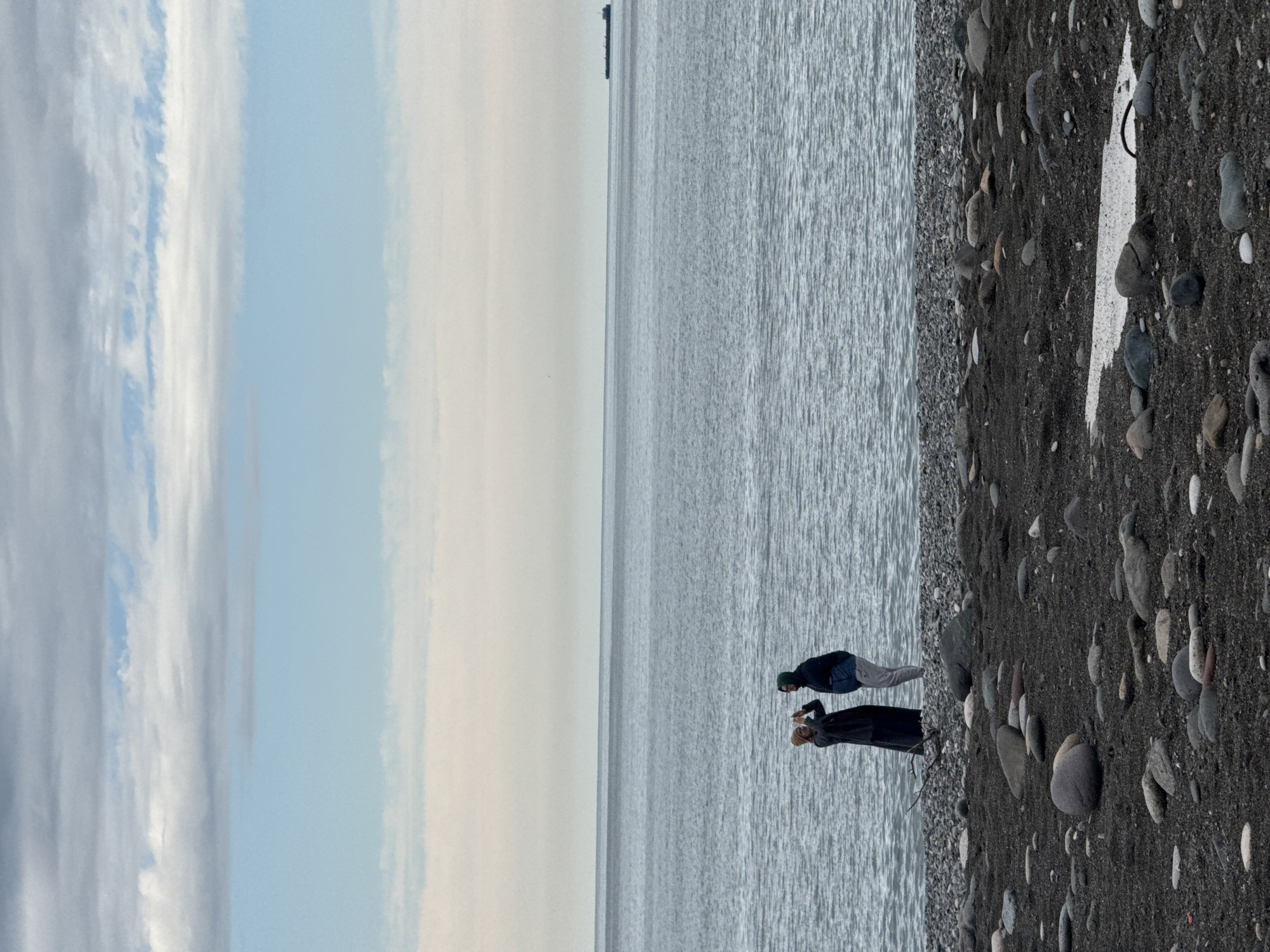 The image depicts a serene seaside scene in Batumi, Georgia, under a soft, diffused sky. The frame is divided horizontally into three main sections: the sky, the sea, and the beach.
The upper portion reveals a partially cloudy sky, with stretches of pale blue interspersed with wispy white and light gray clouds. The horizon line is clearly defined, with a faint haze softening the transition between the sky and the distant water.
The middle section is dominated by the calm, expansive surface of the Black Sea, rendered in cool gray-blue tones. Subtle ripples on the water's surface suggest a gentle breeze. In the far distance, a thin, dark vertical structure, possibly a boat or a marker, is barely visible on the horizon.
The lower third of the image shows the dark, pebbly beach, characteristic of Batumi. The beach is covered in an assortment of smooth, rounded stones of varying sizes and dark hues. Near the water's edge, two individuals are present. One person, dressed in a dark jacket and light pants, possibly wearing a beanie, stands facing the water. Beside them, another person, clad in a long dark garment and a light-colored head covering, is holding up a device, seemingly capturing the scenic view with a phone or small camera.
Notable details on the beach include a distinct white, somewhat triangular mark on the dark pebbles, and a dark, hoop-like object nearby. A small piece of driftwood is also visible near the figures. The overall lighting suggests it's daytime, likely morning or afternoon, with no harsh shadows, contributing to a calm and contemplative atmosphere. There is no visible text in the image.