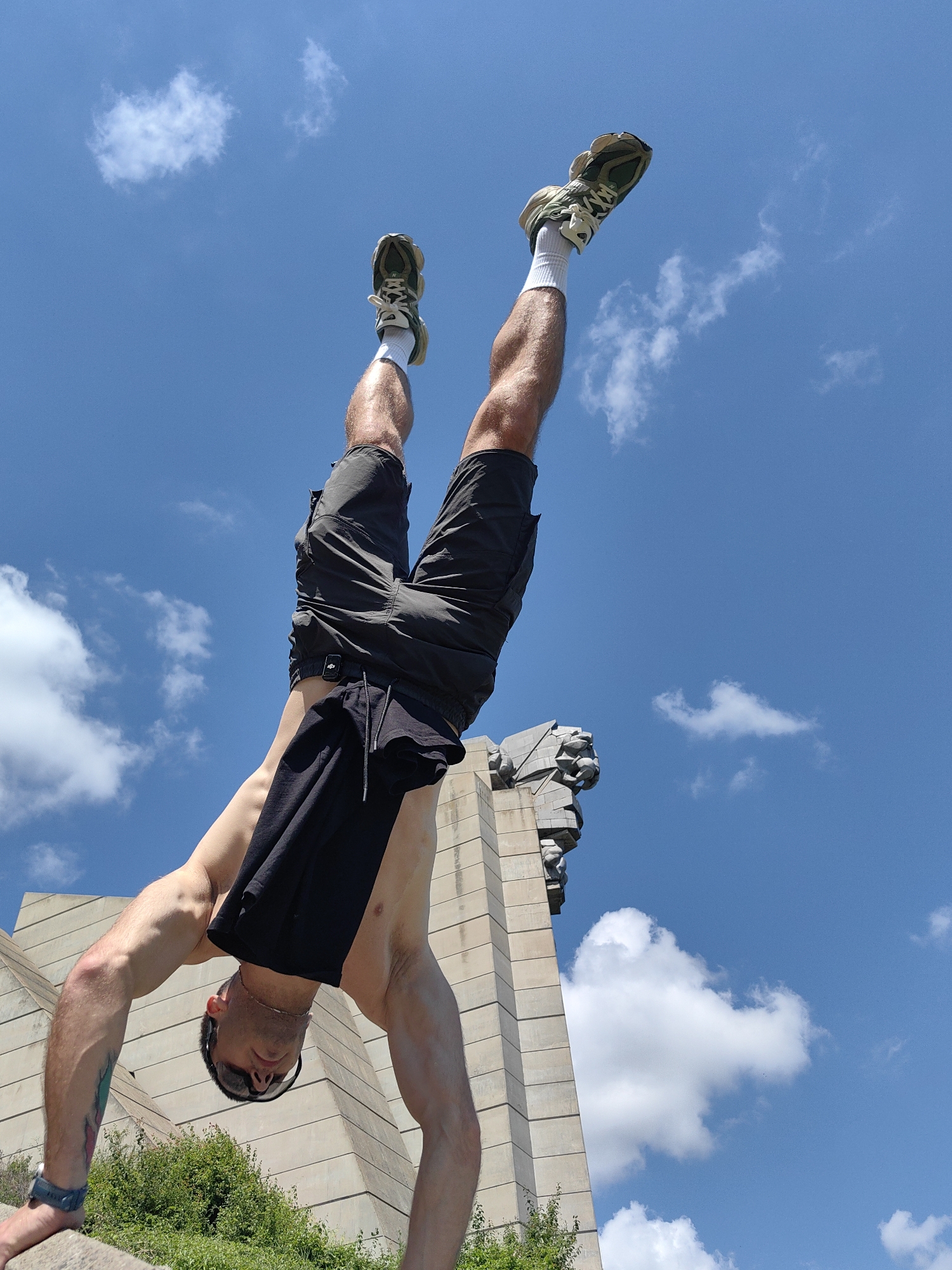 The image shows a man performing a handstand against a bright blue sky with scattered white clouds. He is wearing dark shorts and white socks with athletic shoes. His legs are extended upwards, and a black t-shirt is draped over his waist. In the background, a portion of the Buzludzha Monument, a Brutalist monument located in Shumen, Bulgaria, is visible. The monument is made of concrete and features a large, imposing structure. Green foliage is present at the bottom of the frame, suggesting an outdoor setting. The man's muscles are visible, indicating physical exertion. The perspective is from below, looking up at the man and the monument.