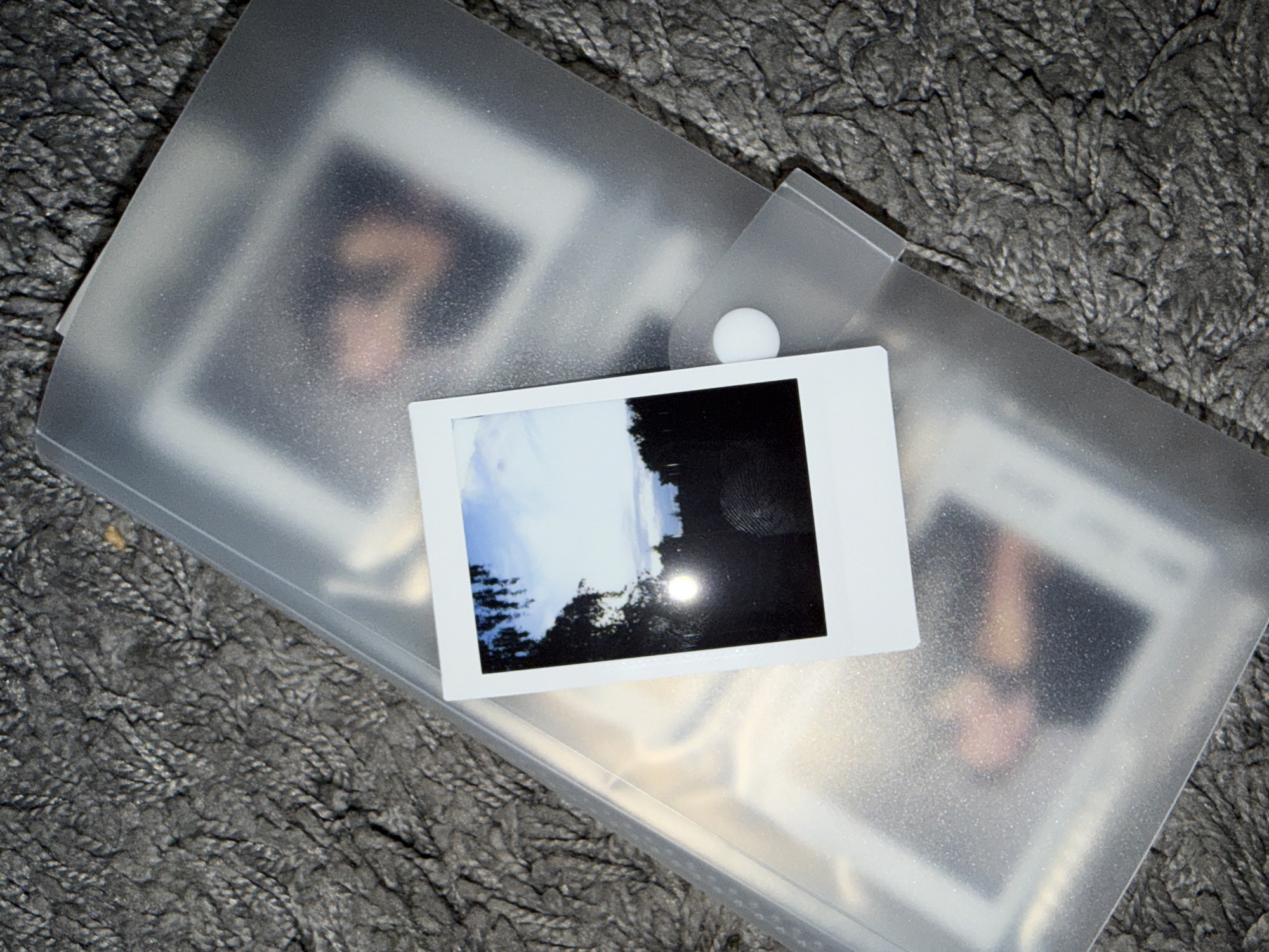A single, slightly unfocused instant photograph is placed on top of a clear plastic folder. The photograph depicts a sunset or sunrise over a dark silhouette of trees and a building. The sun appears as a bright, hazy orb. The plastic folder contains other photographs, which are blurred and indistinct, showing hints of human figures and warm tones. The folder is lying on a textured, grey, carpet-like surface. There is no discernible information within the image to suggest a specific location in Vilnius, Lithuania.