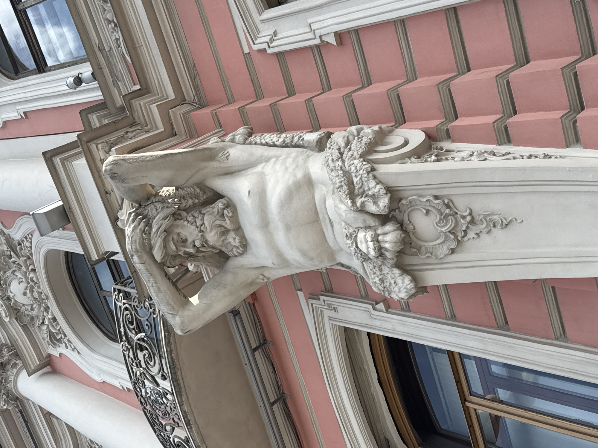 The image shows an exterior architectural detail of a building in Saint Petersburg, Russia. Centered is a sculptural relief of a male figure appearing to support an architectural element. The figure, likely made of stone or plaster, has a detailed facial expression, including a beard and head draped with decorative elements. The background features a pink facade with gray vertical stripes. Windows are visible, reflecting the sky. The decorative trim around the windows and the overall style suggest a baroque or neo-baroque architectural style.