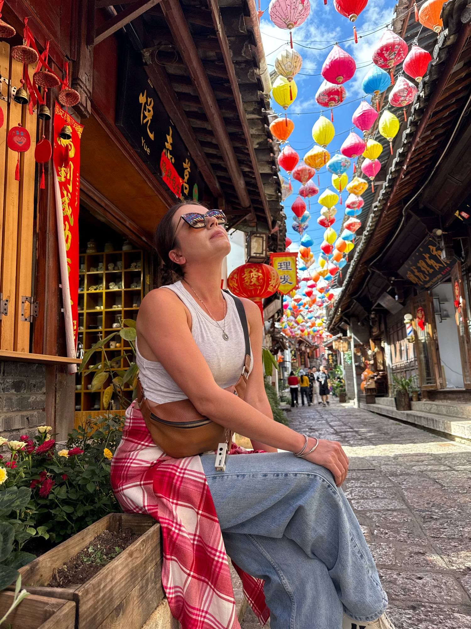 In the Gucheng District of China, a woman sits on a stone bench on a cobblestone street lined with traditional wooden buildings. She is wearing a white tank top, blue jeans, and sunglasses, with a red and white plaid scarf draped over her lap. A brown fanny pack is worn across her chest. Overhead, a multitude of colorful paper lanterns are suspended, creating a vibrant canopy against the blue sky. The street is bustling with activity, with people walking and shops lining both sides. The architecture is distinct, featuring dark wood eaves and ornate signage in Chinese characters. The woman is looking upwards, seemingly captivated by the lanterns. The foreground features a wooden planter box filled with flowering plants.