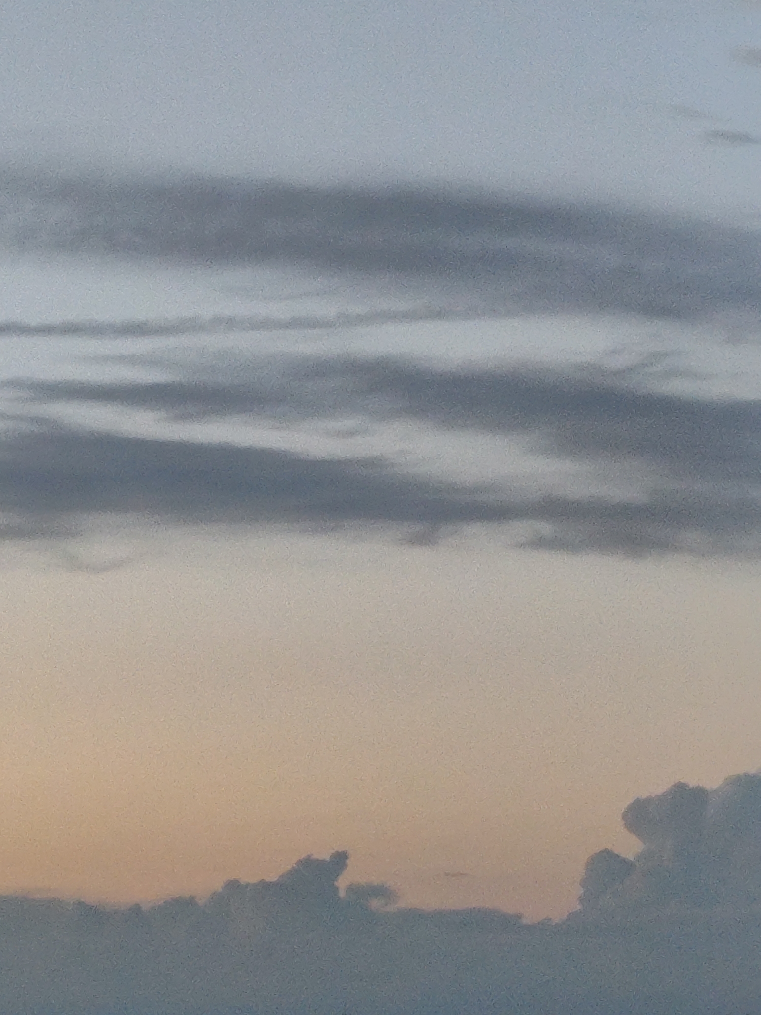 The image depicts a sky with layers of clouds. The upper portion of the image is a pale blue-gray, transitioning to a warm orange-peach hue closer to the horizon. Multiple bands of darker gray clouds are visible horizontally across the sky, some appearing wispy and others more solid. At the very bottom of the image, a darker, more defined cloud formation is visible, suggesting a landscape or horizon line. The scene appears to be a natural landscape, possibly a sunset or sunrise. The image does not contain any identifiable landmarks or features that would indicate its specific location within Zielona Góra, Poland. There are no people, animals, or specific objects clearly visible in the frame.