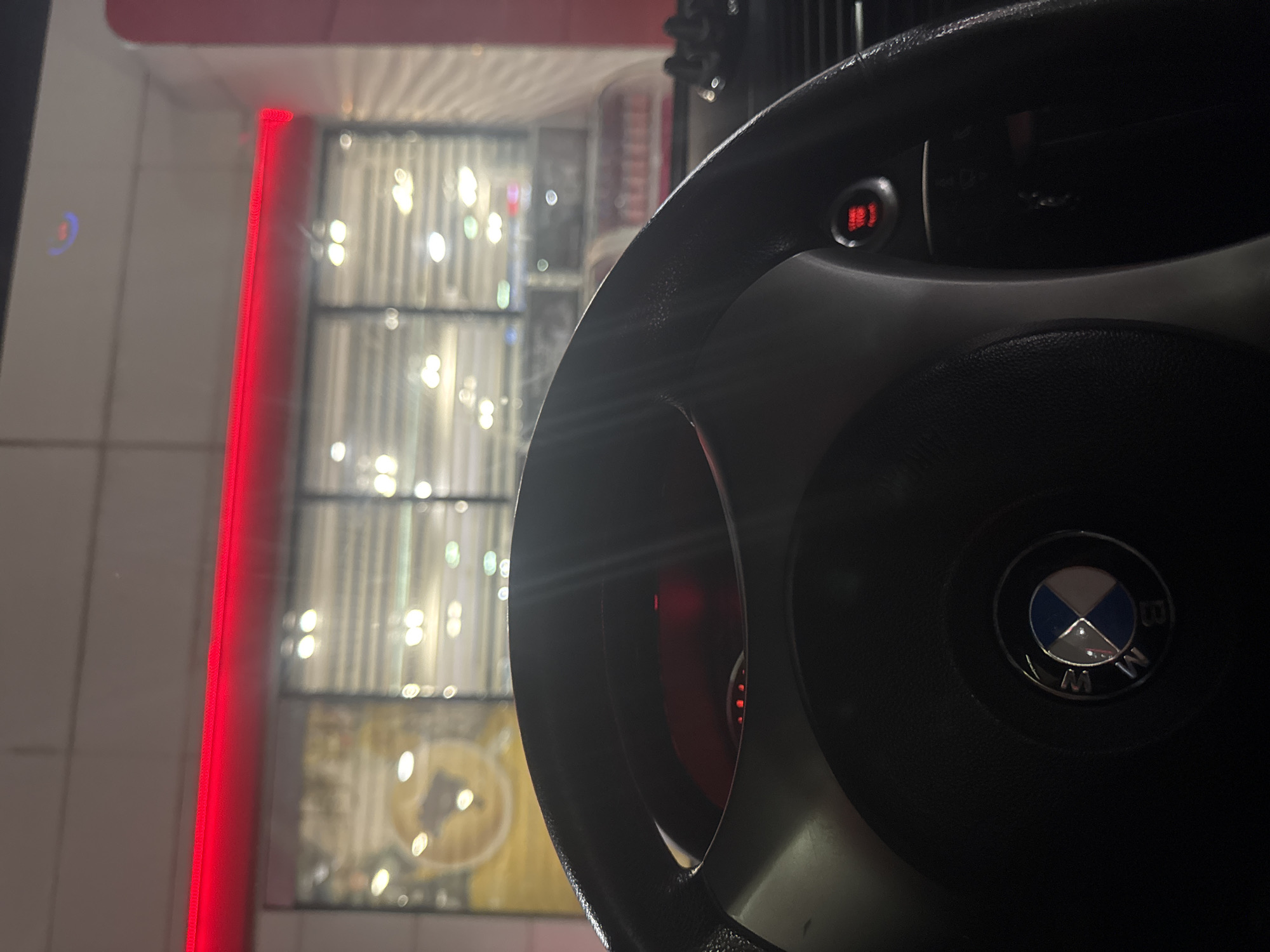A close-up photograph shows the steering wheel of a BMW car. The steering wheel is black and features the BMW logo in the center. The background reveals a building with a red neon light outlining its edge and windows with illuminated displays. The blurred lights suggest an urban environment, potentially a store or business establishment. While the location is noted as Vodenichane, Bulgaria, specific contextual details beyond what is visible in the image cannot be determined.