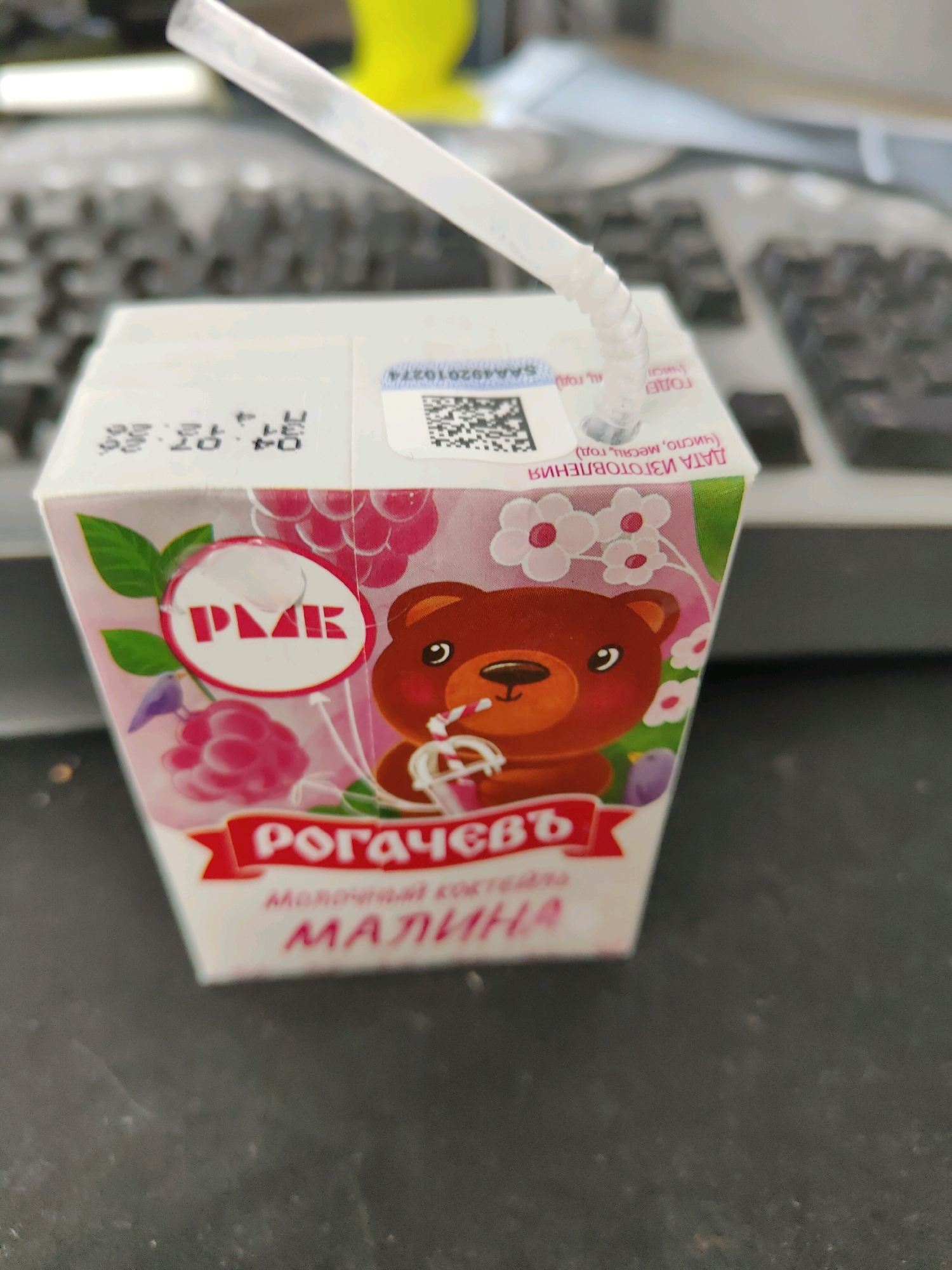 A close-up, eye-level shot shows a juice box with a straw inserted into the top, resting on a dark surface, possibly a desk or table, with a keyboard visible in the blurred background. The juice box is white with a colorful illustration featuring a cartoon brown bear, raspberries, flowers, and a small bird. The brand name "РОГАЧЕВЪ" is prominently displayed in red text on a white banner across the lower portion of the juice box, followed by "МОЛОЧНЫЙ КОКТЕЙЛЬ" (milk cocktail) and "МАЛИНА" (raspberry) in pink text. There are also some markings and a barcode on the top of the box. The lighting appears to be overhead, creating some reflections on the glossy surface of the juice box. The overall scene is a still life, with no people or overt actions captured. The image does not provide any direct or indirect information about the location in Homyel, Belarus.