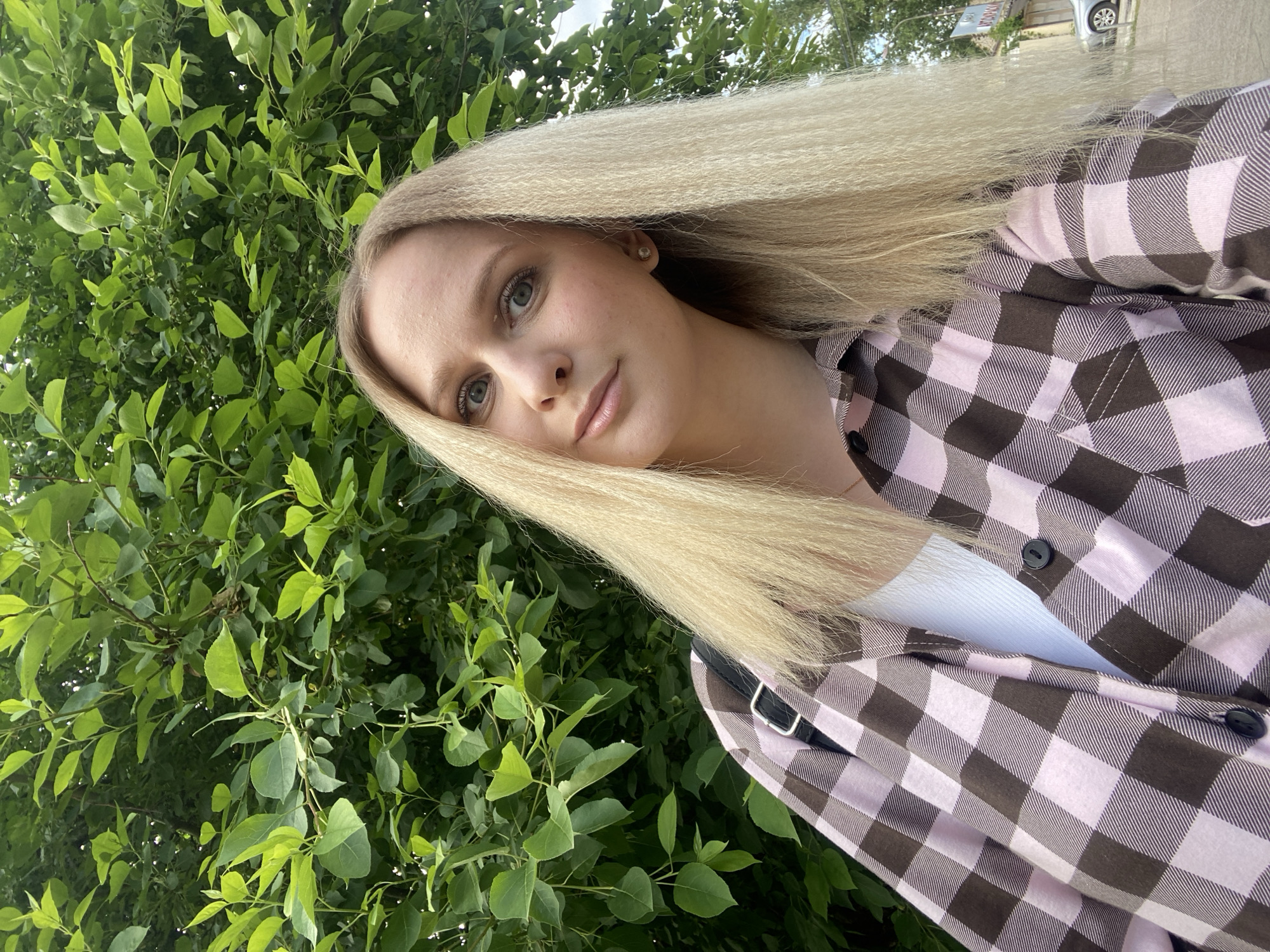 A portrait-style selfie captures a young woman outdoors in Chaikovsky, Russia. She has long, very light blonde hair with a distinct crimped texture, fair skin, and light-colored eyes. She looks directly at the camera with a calm and gentle expression. She is wearing a long-sleeved button-up shirt featuring a prominent light pink and dark brown checkered pattern, layered over a white top. A small, light-colored stud earring is visible in her right ear, and a black strap, possibly from a bag, crosses her right shoulder.
The background is dominated by lush green foliage from trees or bushes, indicating a natural outdoor setting during daytime with bright, natural light. The image is tilted, creating a dynamic and informal perspective. In the upper right corner of the background, partially obscured by leaves, a blurred white car and an indecipherable sign are faintly visible, suggesting proximity to a road or developed area. The only activity depicted is the woman posing for the photograph. No specific text is readable within the image.