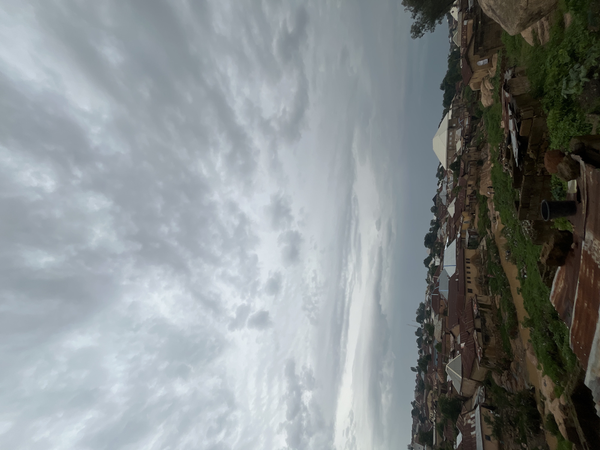 The image captures a view of a densely populated hillside settlement, likely in Jos, Nigeria. The scene is dominated by a sky filled with overcast, grey clouds, suggesting either early morning, late evening, or a day of impending rain. The settlement itself is characterized by numerous buildings with corrugated iron roofs, built closely together along the undulating terrain. Many of the structures appear to be made of brick or mud, reflecting traditional building methods. Green vegetation is visible in patches, clinging to the hillsides and in between the houses. In the foreground, there are some signs of disrepair and rustic elements, including what appear to be discarded metal objects or debris, adding to the raw, lived-in feel of the location. A large, modern building with a white, pitched roof stands out among the more traditional structures, possibly a school, community center, or religious building. The orientation of the image is vertical, presenting the hillside and the sky in a landscape format. There are no discernible people or specific activities taking place, but the general impression is one of a bustling, if somewhat rugged, urban environment shaped by its geography. The overall mood is subdued, reflecting the cloudy weather and the dense, layered architecture.