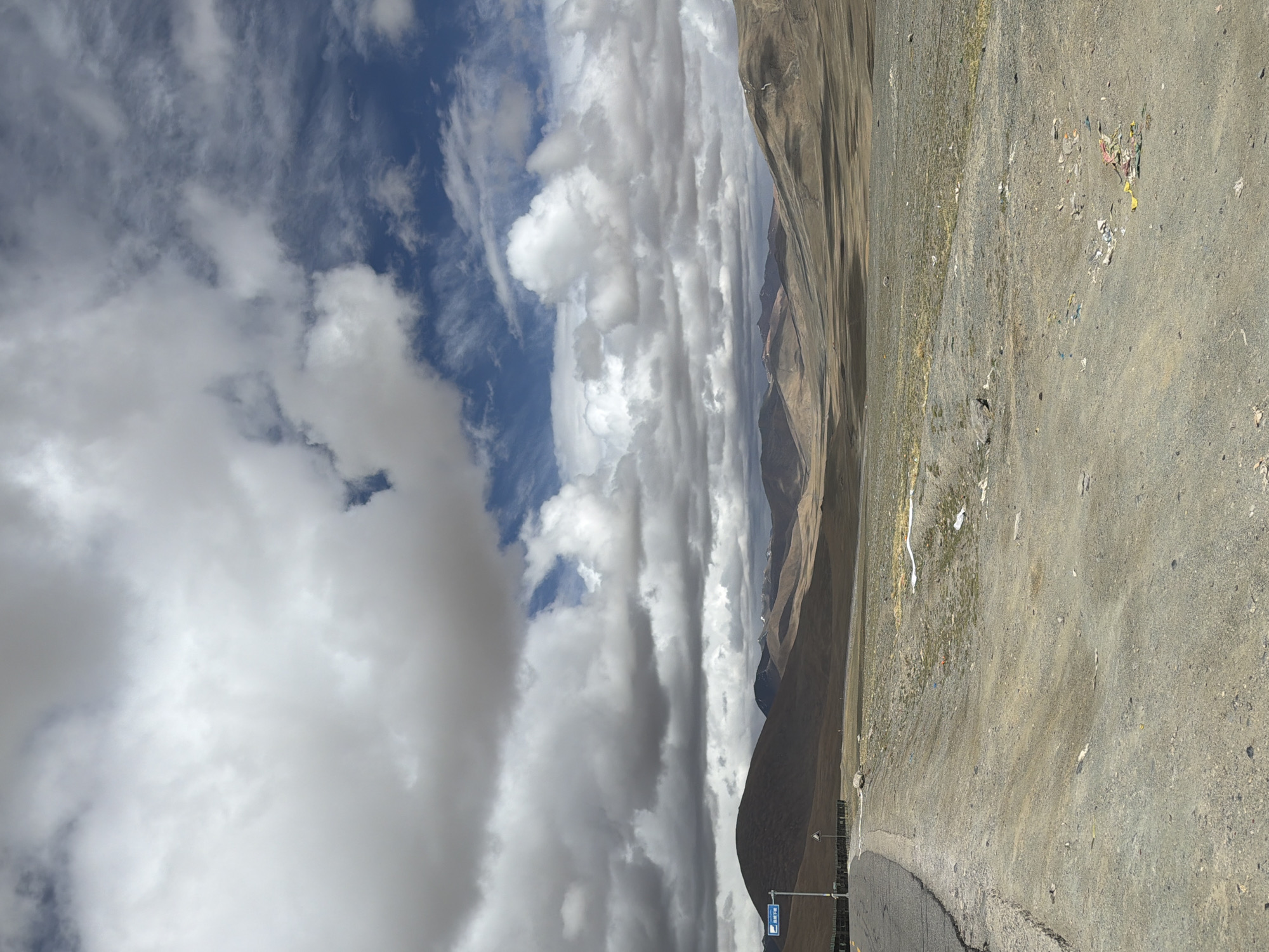 The image depicts a landscape near Hor Town in China, under a sky filled with dramatic clouds. The mountains appear in the background, showing a range of brown and grey tones. A road runs along the right side of the frame, and the ground adjacent to the road is rocky and barren. There are some scattered prayer flags along the roadside. A sign with Chinese characters is visible near the road.