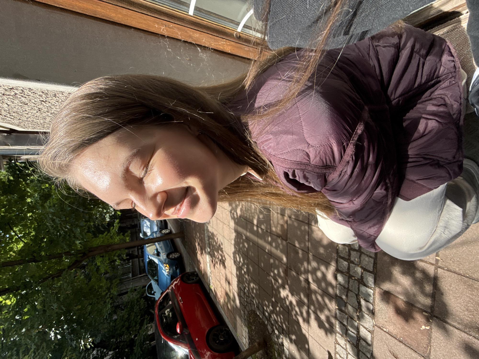 A woman is standing on a brick sidewalk in Belgrade, Serbia. She is wearing a maroon quilted jacket, grey pants, and a white shirt. She is smiling and looking towards the light. Behind her, on the street, there are parked cars, including a red one and two blue ones. Trees line the street, casting shadows on the sidewalk. A building with a window is visible on the left side of the frame.