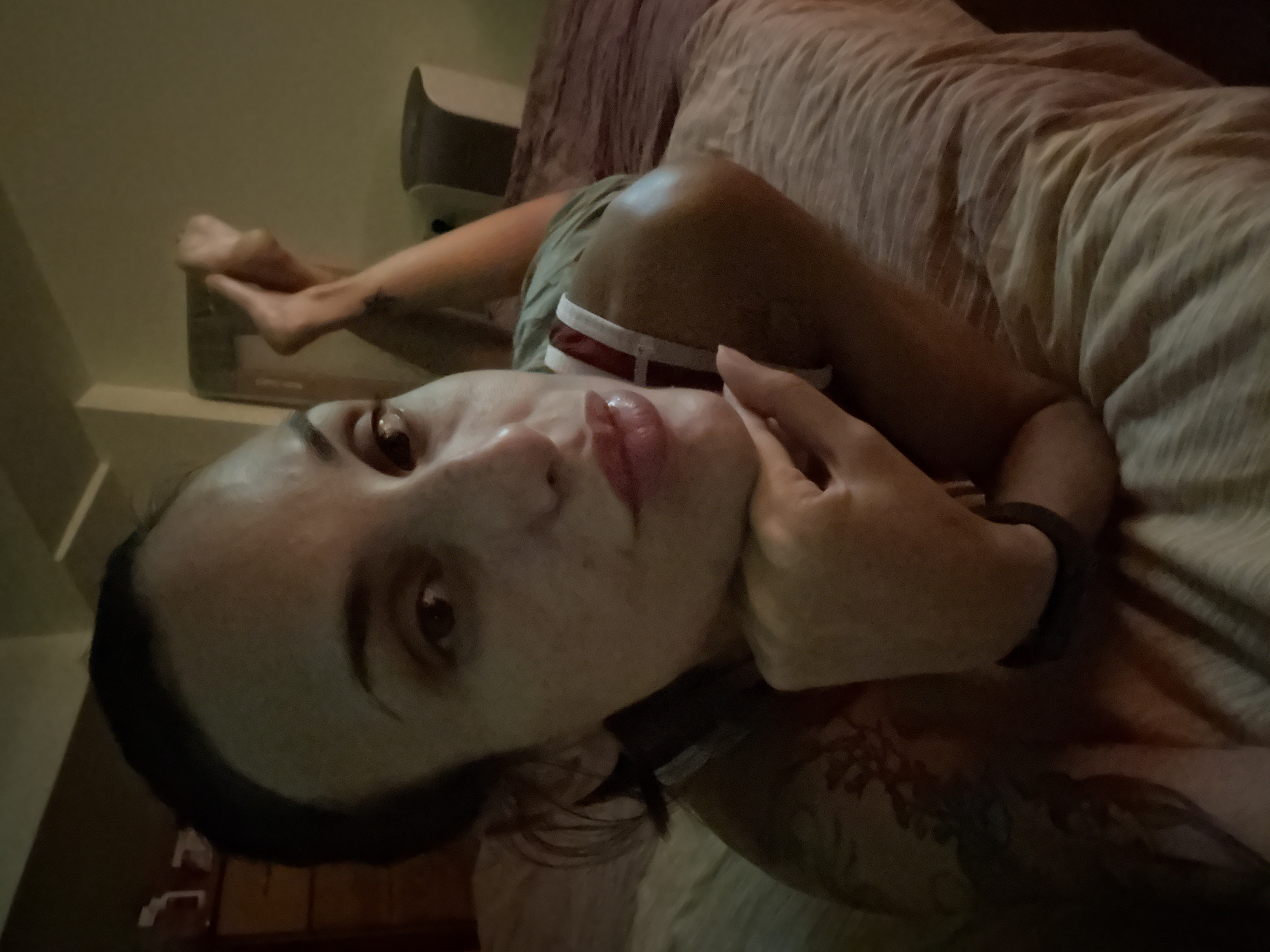 An overhead, close-up shot shows a woman laying on her stomach on a bed. She is facing the camera and resting her head on her hands, with her elbows on the bed. Her legs are bent and her feet are in the air behind her. She is wearing a light-colored shirt with red and white straps. The background is dimly lit and shows a wall and part of a dresser. There are no identifying landmarks of Pleven, Bulgaria visible in the image.