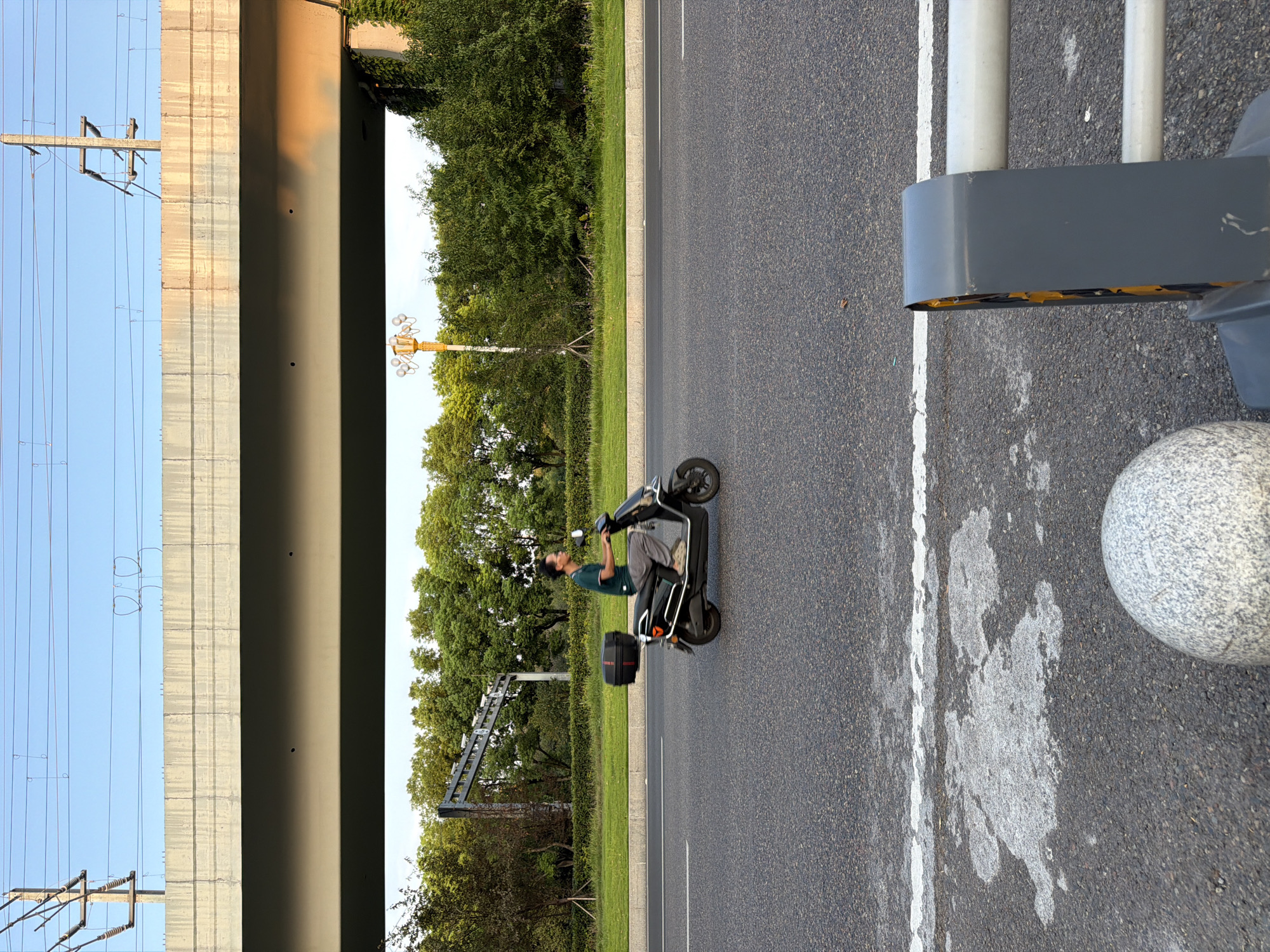 In Jiangjin, China, a man rides a black electric scooter on a paved road. The scooter has a black storage box mounted on the rear. The man is wearing a dark green t-shirt and light-colored pants. He is looking forward. The road is a multi-lane asphalt surface with white lane markings. In the background, there is a large concrete overpass with visible electrical infrastructure above it. Lush green trees and vegetation line the side of the road. The sky is clear and blue with the soft glow of the setting sun reflecting on the overpass. In the foreground, a portion of a gray barrier and a speckled stone sphere are visible.