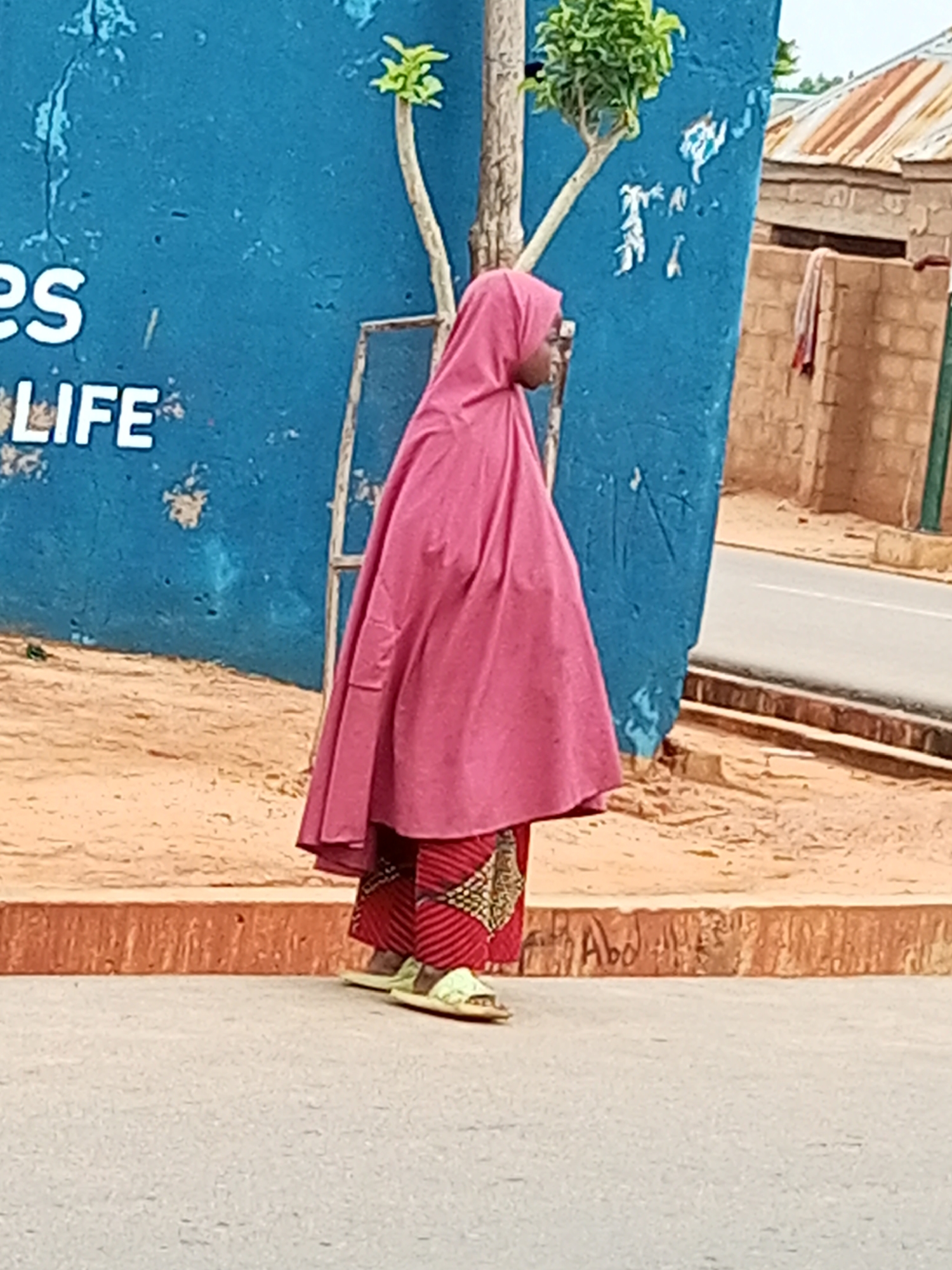 A full-shot, side-profile image captures a person wearing a pink hijab and a patterned red and gold dress, standing on a sandy area next to a bright blue wall with white lettering that reads "LIFE." The person is looking to the left, with their feet clad in light-colored sandals. The wall is textured and peeling in places, with a thin tree growing beside it. Beyond the wall, a dusty road is visible, with buildings constructed from brick and corrugated metal roofing in the background. The lighting suggests it is daytime. The scene appears to be located in Tempure, Nigeria.