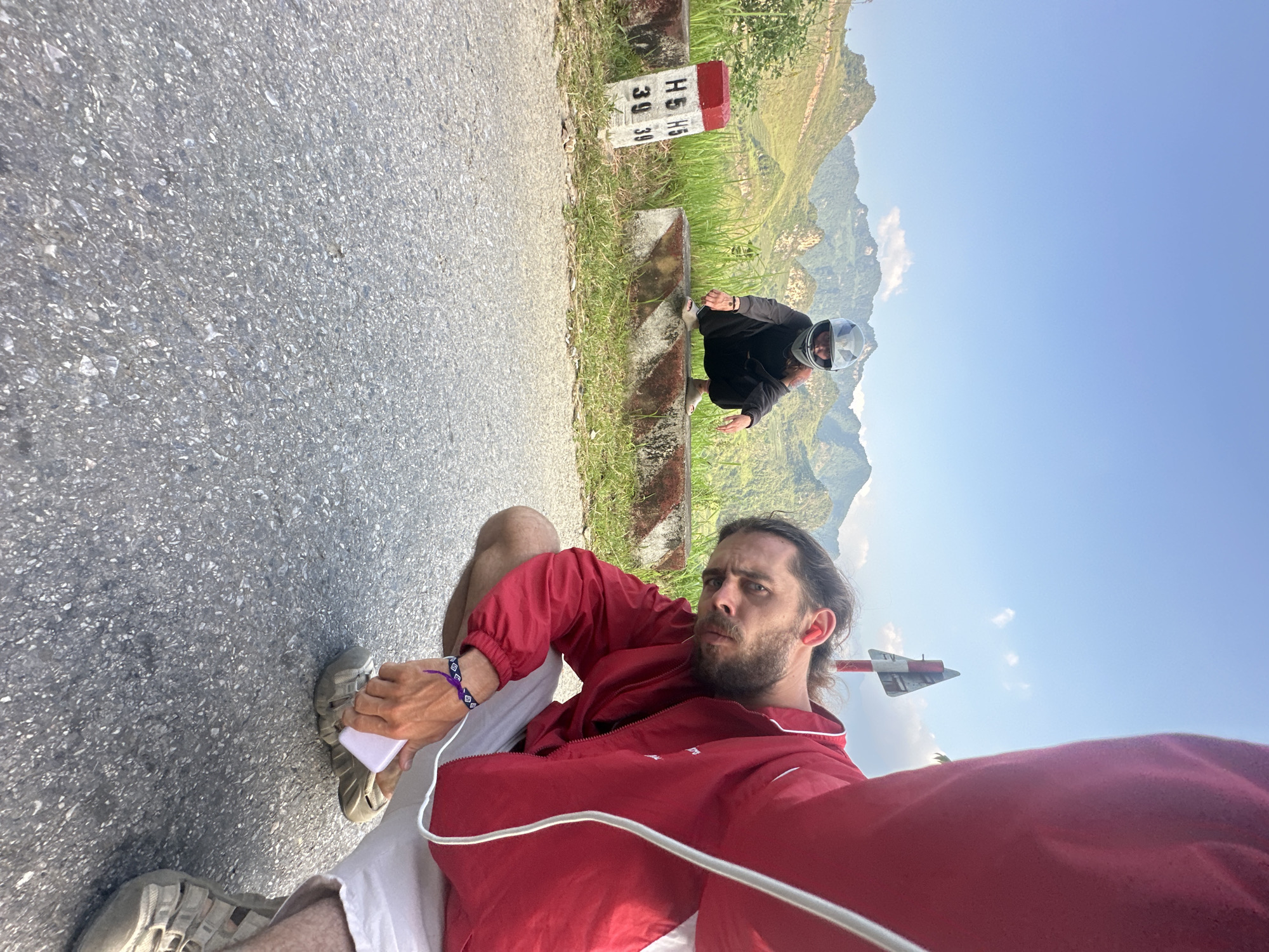 A man in a red jacket sits on an asphalt road in Du Già Commune, Vietnam, taking a selfie. In the background, another person wearing a motorcycle helmet squats near a roadside marker post displaying numbers "H5" and "39" on its sides. The landscape features mountains under a clear sky. The immediate foreground is dominated by the road surface texture and the man's clothing and footwear.