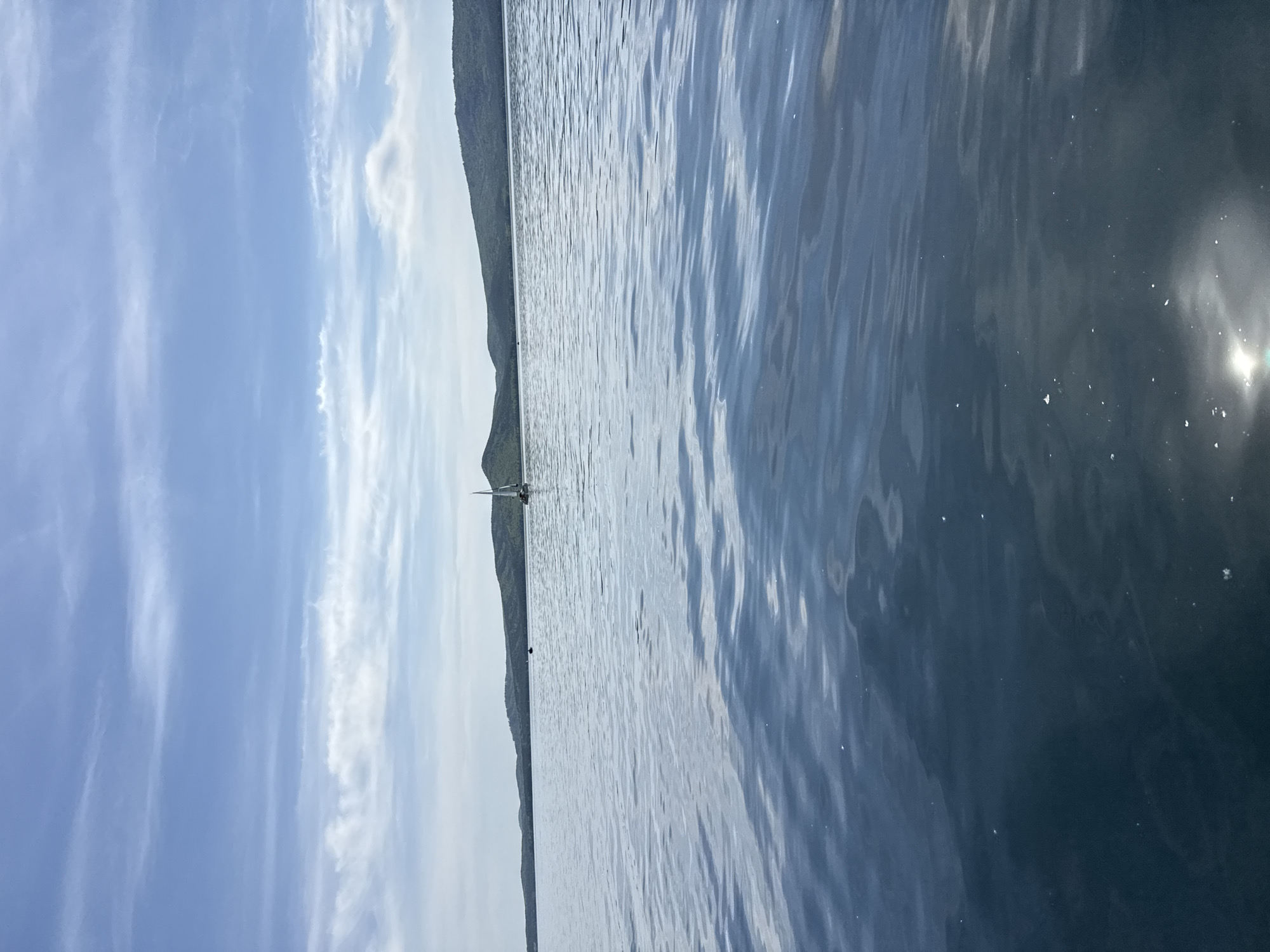 The image shows a body of water with gentle ripples on the surface, reflecting sunlight. In the distance, a shoreline is visible with green hills or mountains. A sailboat is positioned on the water near the shoreline. The sky is blue with wispy white clouds. The location is likely near Zolotoi Plyazh, Russia, judging by the landscape.