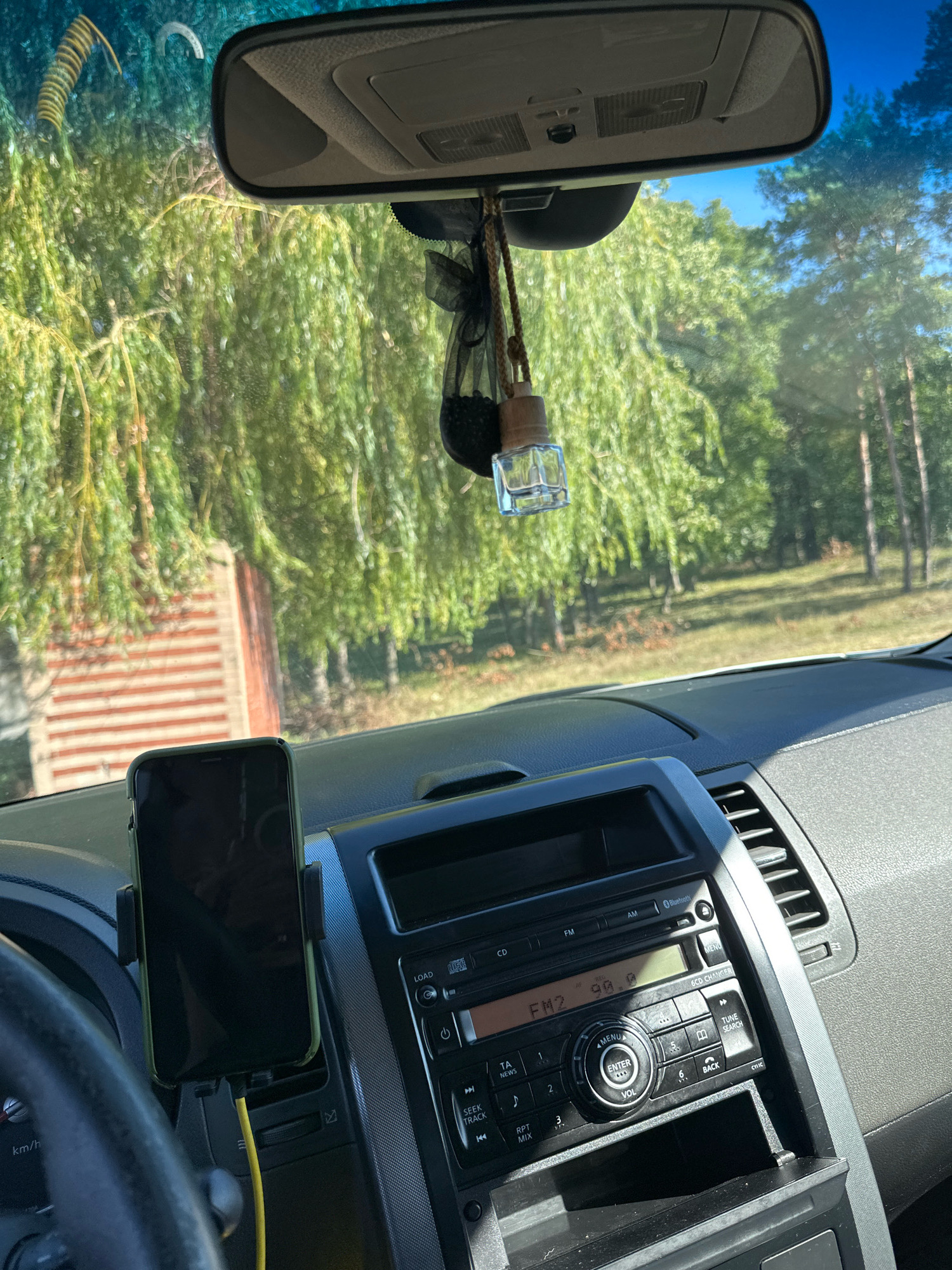 The image shows the interior of a car, looking out through the windshield. Through the windshield, we see trees and a blue sky. Inside the car, we can see the dashboard with a car stereo, a phone mounted on a vent, and a rear-view mirror hanging from the ceiling. A car air freshener is hanging from the rear-view mirror. The location is indicated as Makiv, Ukraine.