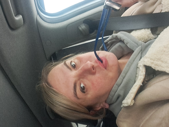 A woman with blonde hair and light complexion is looking up at the camera. She wears a light gray hooded sweatshirt under a beige jacket with a fluffy collar. A blue tube protrudes from her mouth. She is seated in a vehicle with a gray interior, wearing a seatbelt. A window is visible in the background.