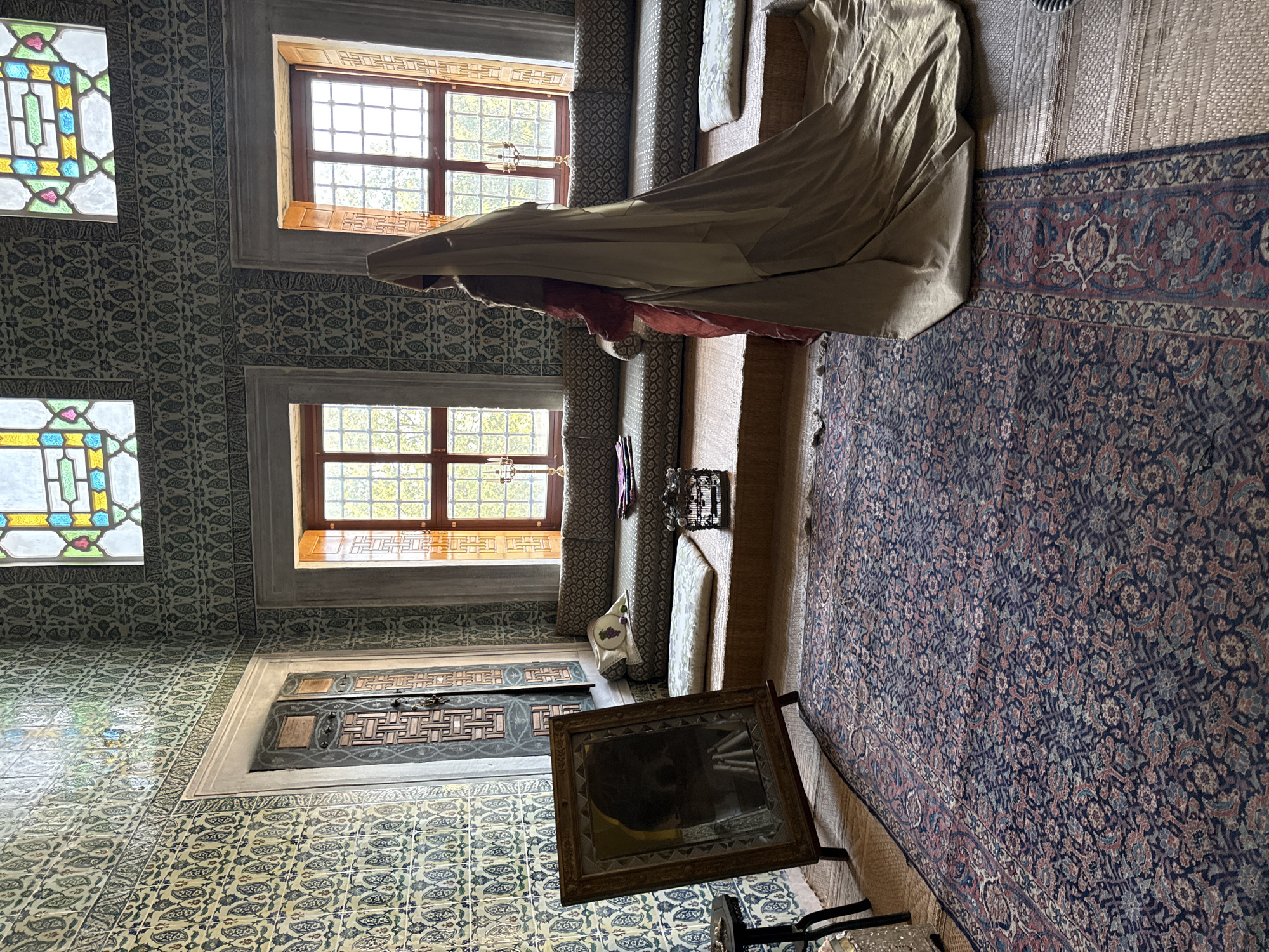 The image depicts an ornate room within the Topkapi Palace in Istanbul, Turkey, likely a former royal chamber or a recreated historical scene. A woman, dressed in historical attire consisting of a red garment and a flowing beige veil and train, stands facing away from the camera, towards a window. The room is richly decorated with blue and white Iznik tiles covering the walls. Two large windows with wooden frames and grilled panes are visible, one with stained glass accents at the top, letting in natural light.
The floor is covered by a large, intricately patterned oriental rug in shades of blue, red, and beige. Beside the rug, to the right, is a raised platform with cushions and a patterned sofa. A framed mirror on an easel stands to the left of the woman, reflecting a shadowy figure. A small, dark metal birdcage sits on a cushion near the sofa. A small table with a lamp is partially visible on the far left. The atmosphere suggests a preserved historical space or a staged representation of one.